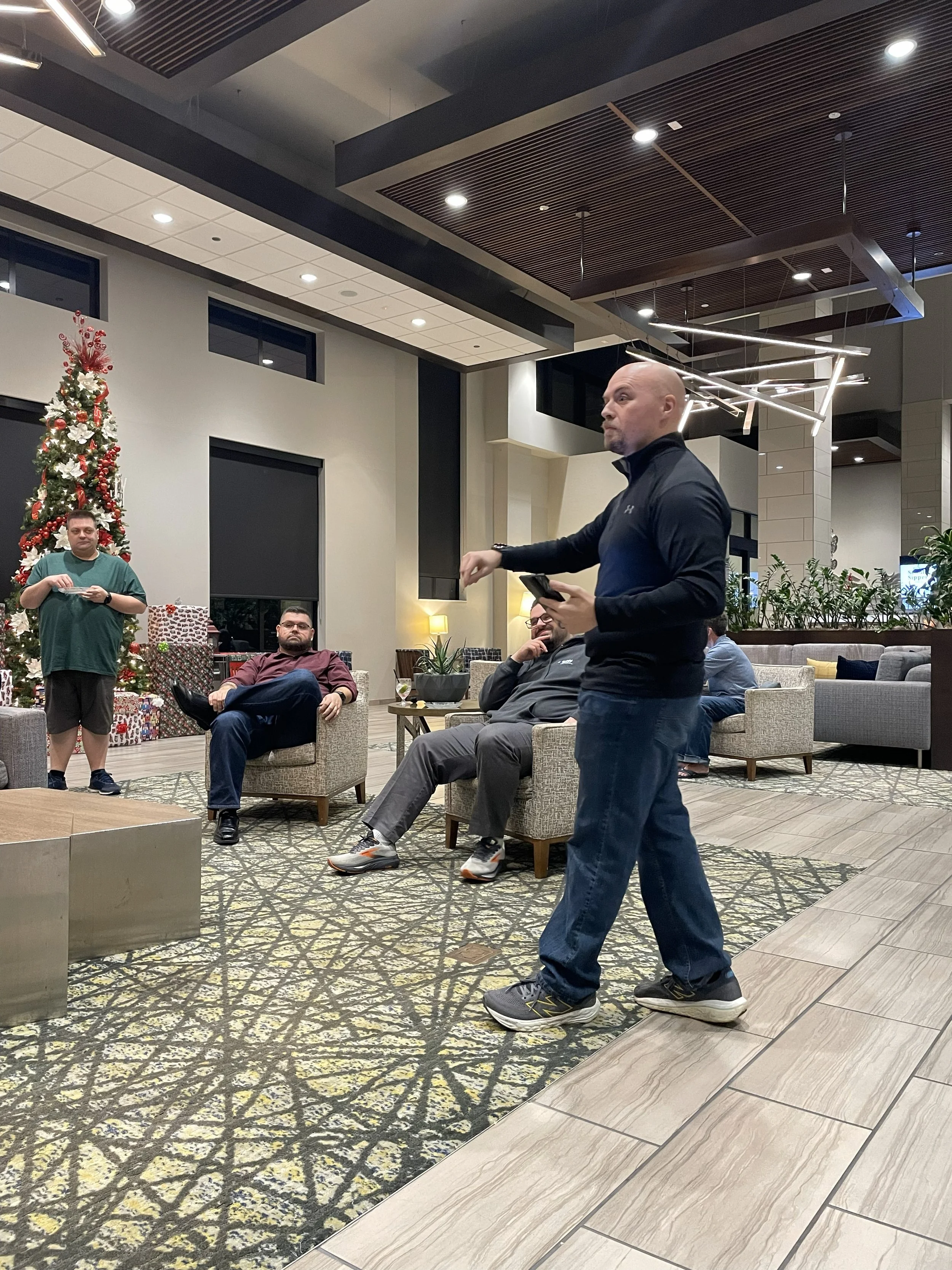 A man in a black and blue athletic tracksuit standing and speaking in a lobby or lounge area with seated people, decorated for Christmas with a large decorated Christmas tree and wrapped gifts, modern lighting fixtures, and a high ceiling.