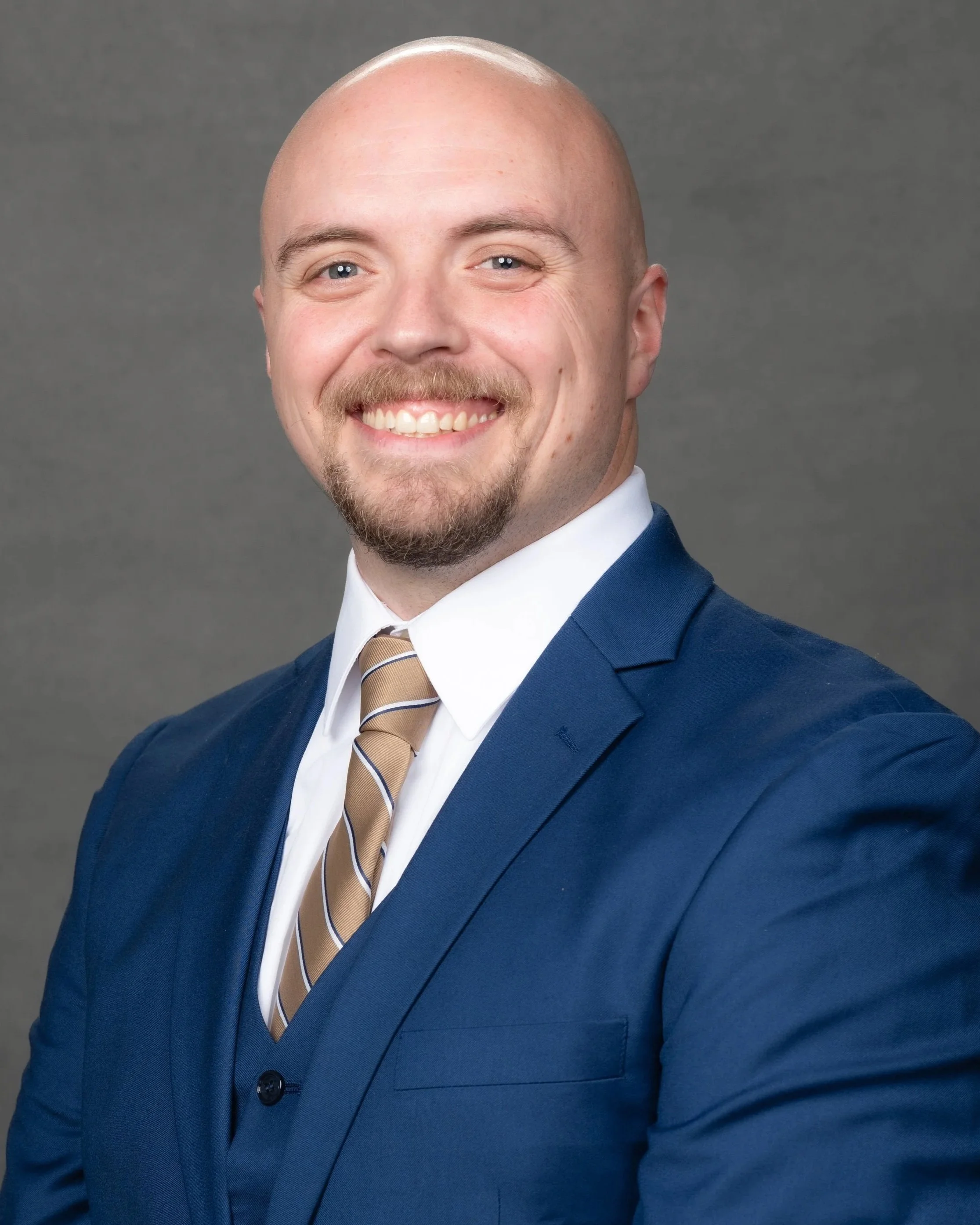 Balding man with a goatee wearing a blue suit, white shirt, and tan striped tie, smiling against a gray background.