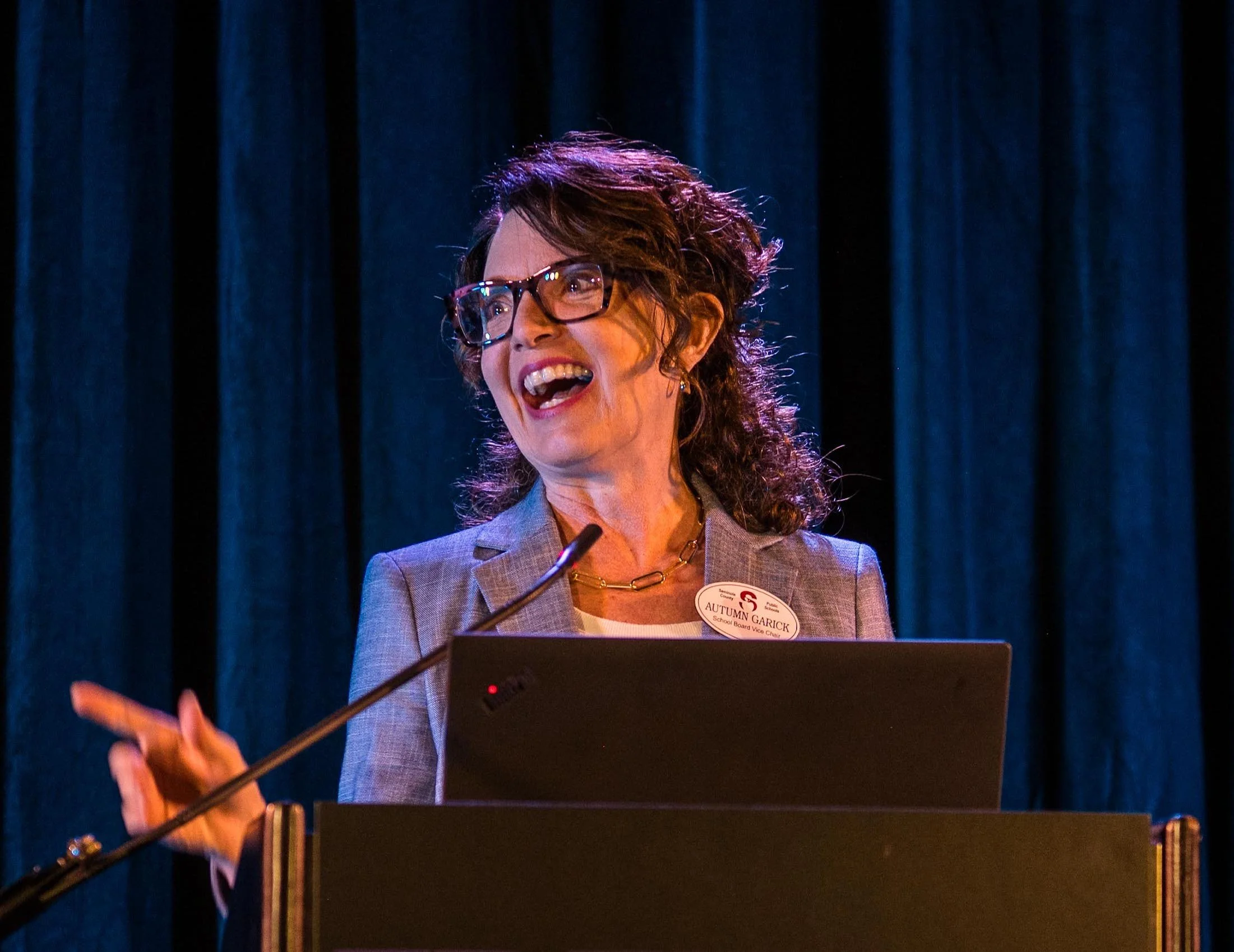 A woman with curly brown hair and glasses, smiling and speaking at a podium with a microphone, against a dark blue curtain backdrop.