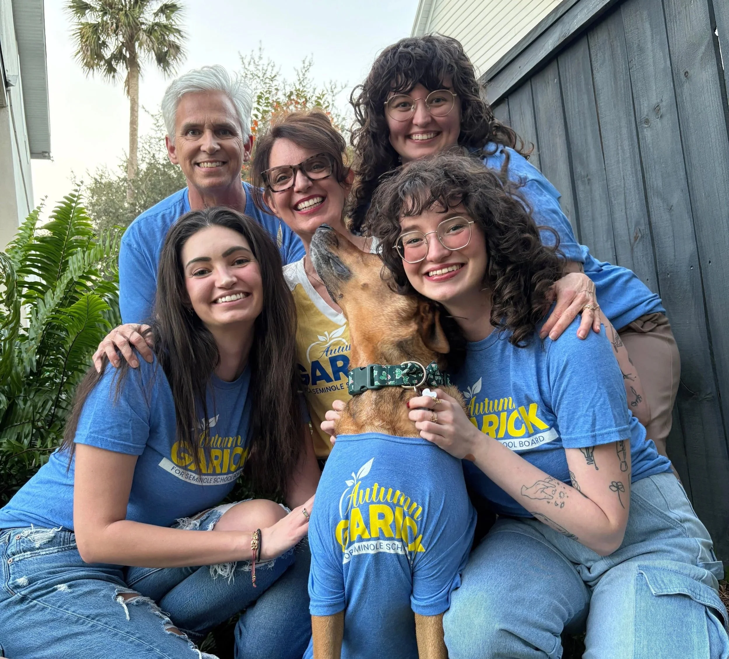 Group of six people and a dog outdoors, smiling and posing for a photo. The dog is wearing a blue shirt with yellow text, and one woman is holding the dog.