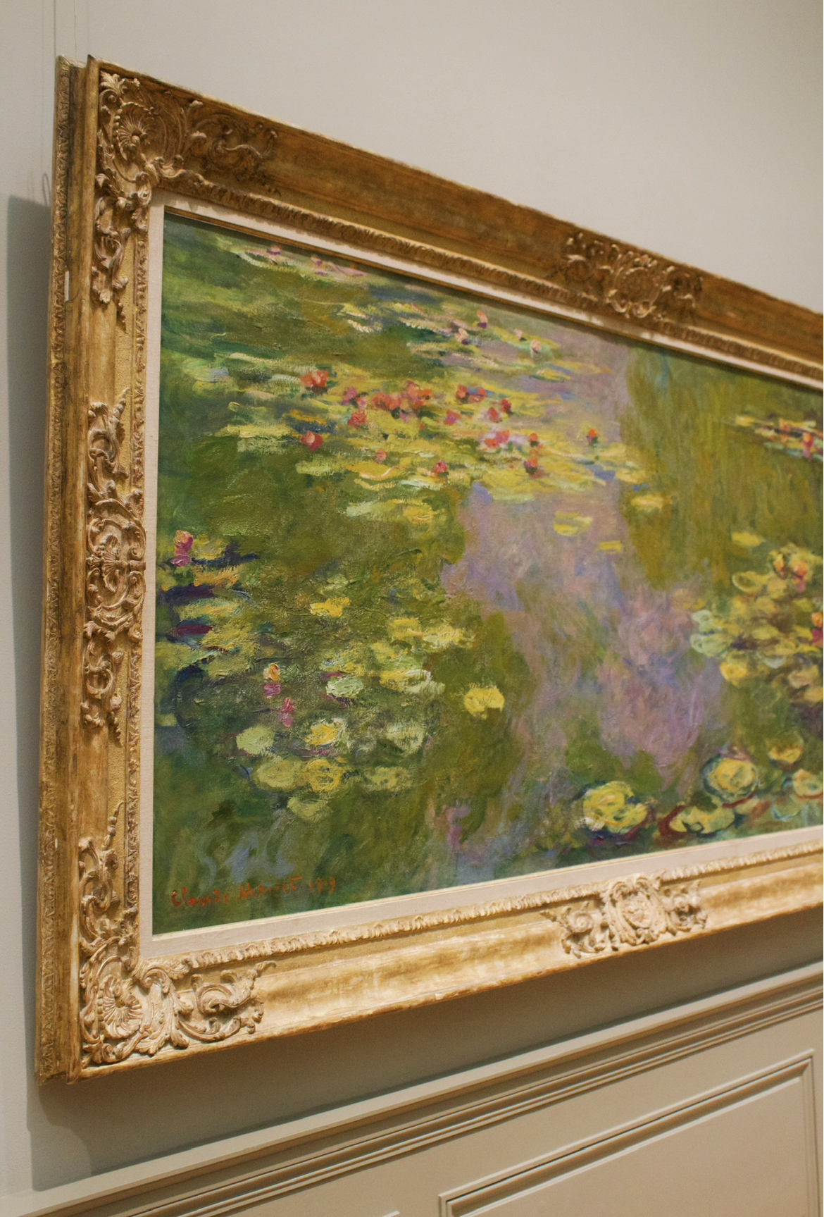 A framed Impressionist-style painting of a pond with water lilies, featuring green, yellow, pink, and purple colors, signed 'Claude Monet 19'.