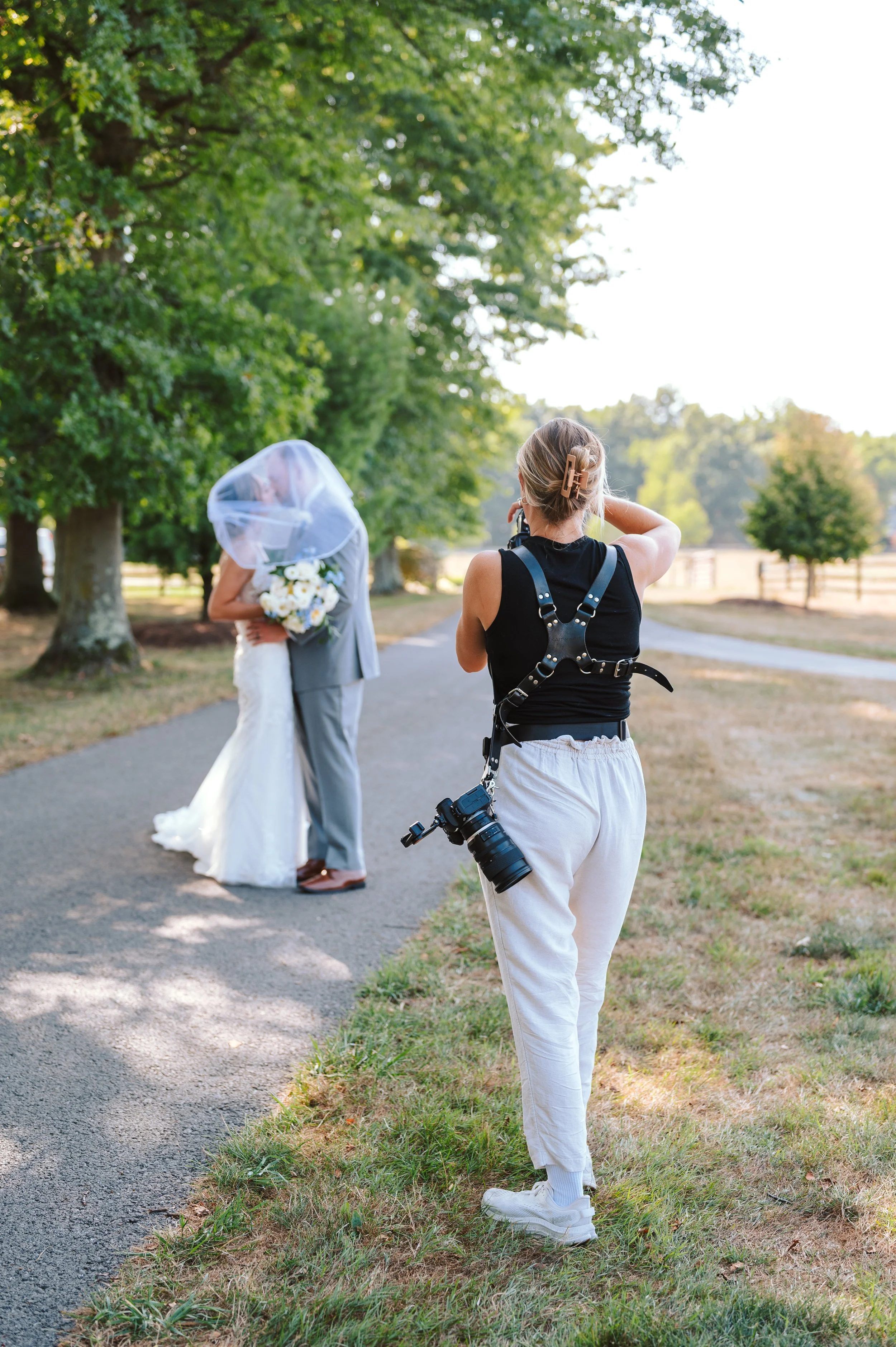 A photographer taking pictures of a couple hugging on a path in a park with trees and fencing in the background.
