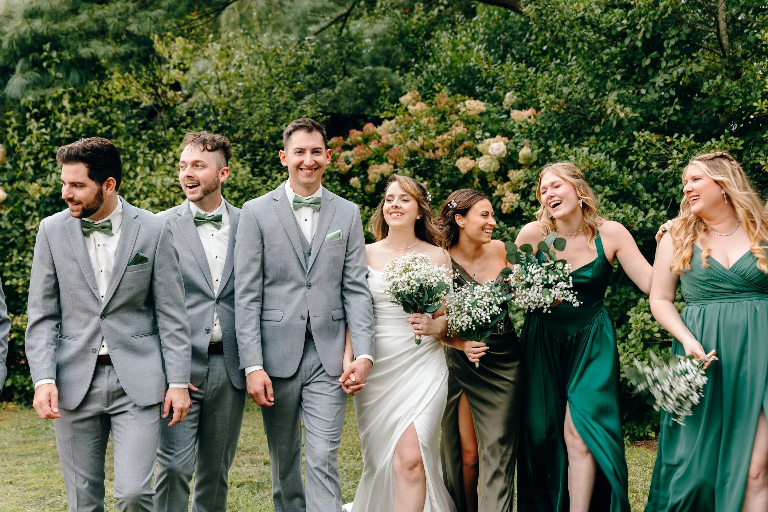 Wedding party walking outdoors amid lush greenery and blooming flowers, dressed in formal attire.