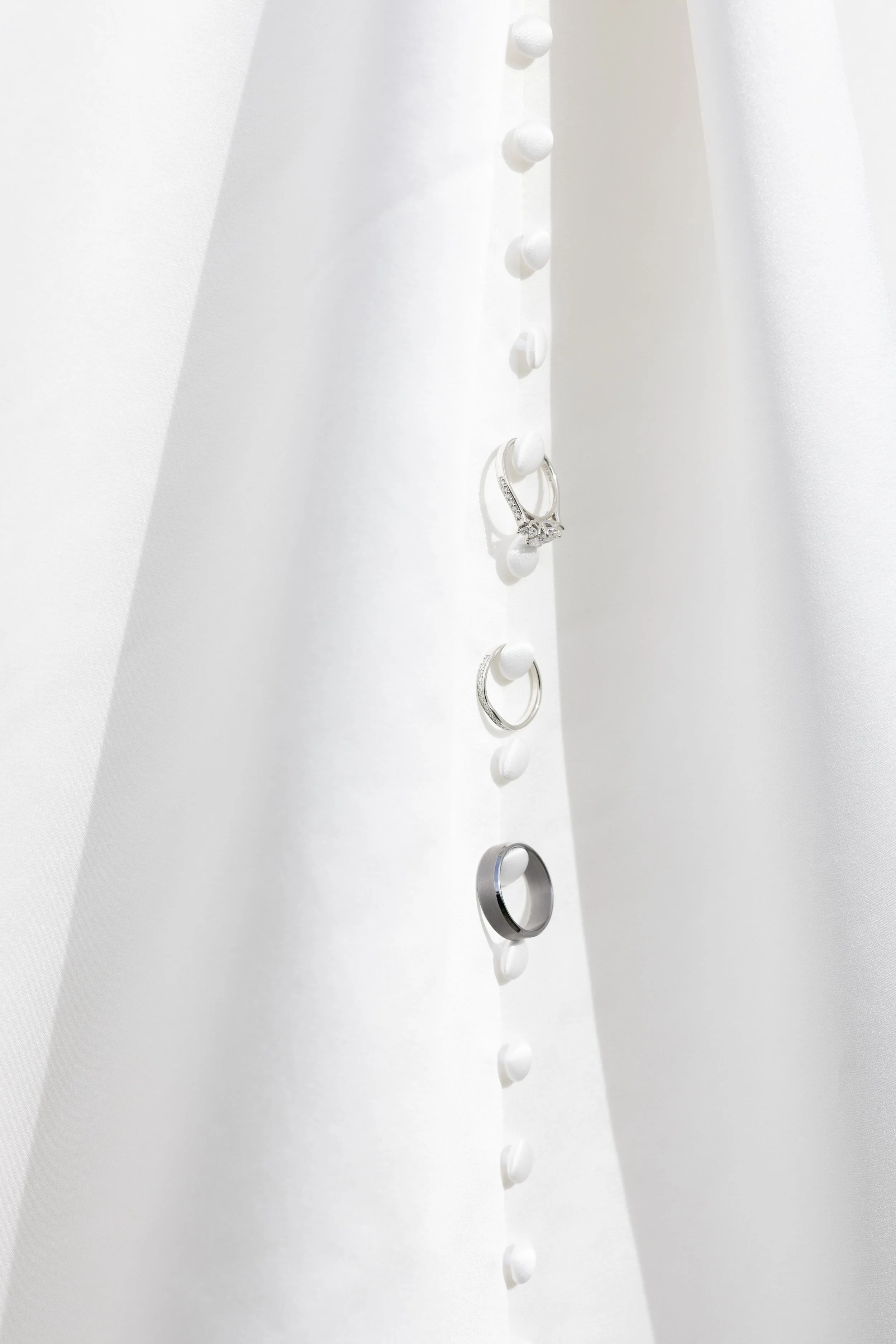 Three silver rings displayed on small white pegs attached to a white fabric background, with soft lighting creating subtle shadows.