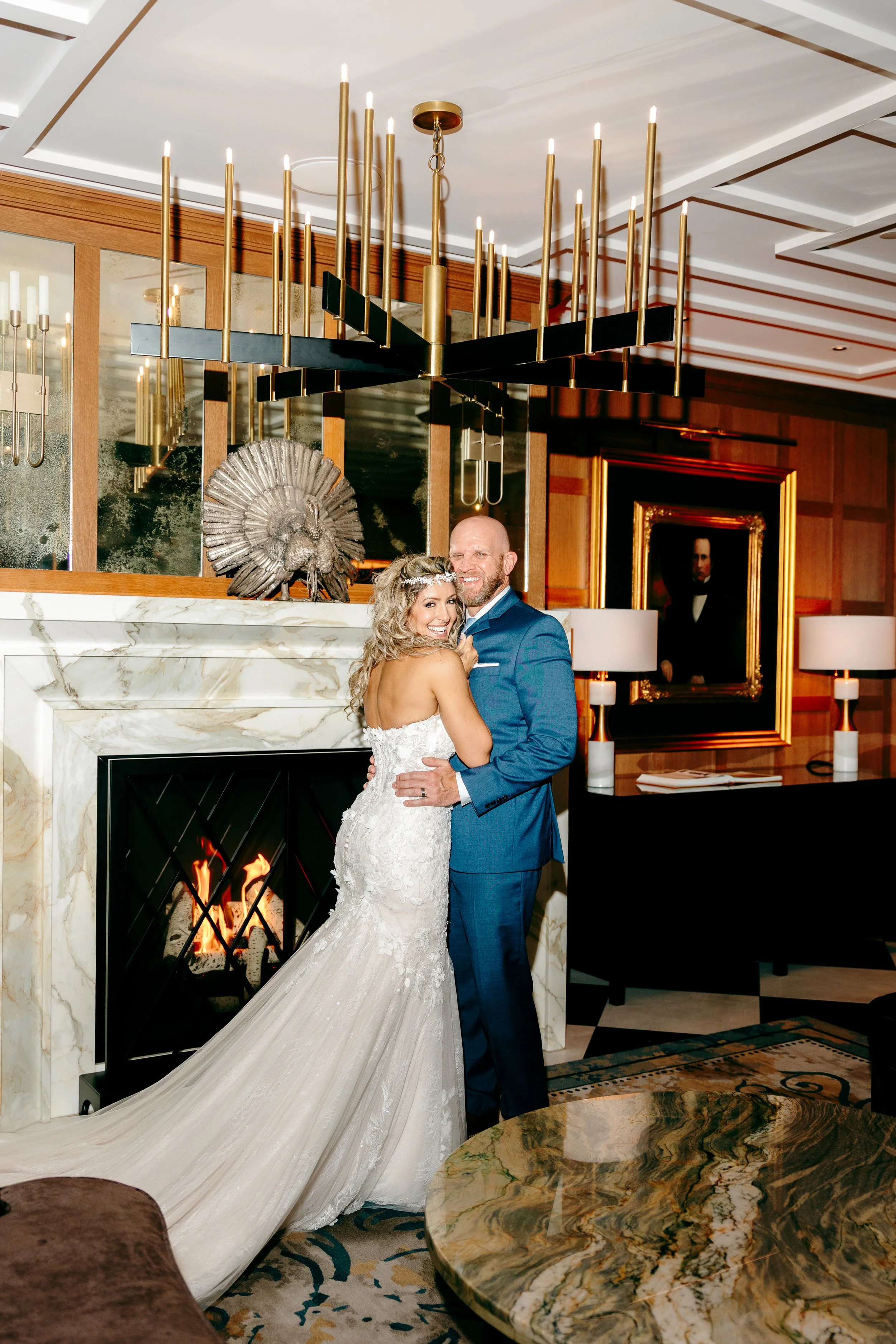 A bride and groom hug each other in a luxurious room with a marble fireplace, a peacock sculpture, and elegant decor, celebrating their wedding.