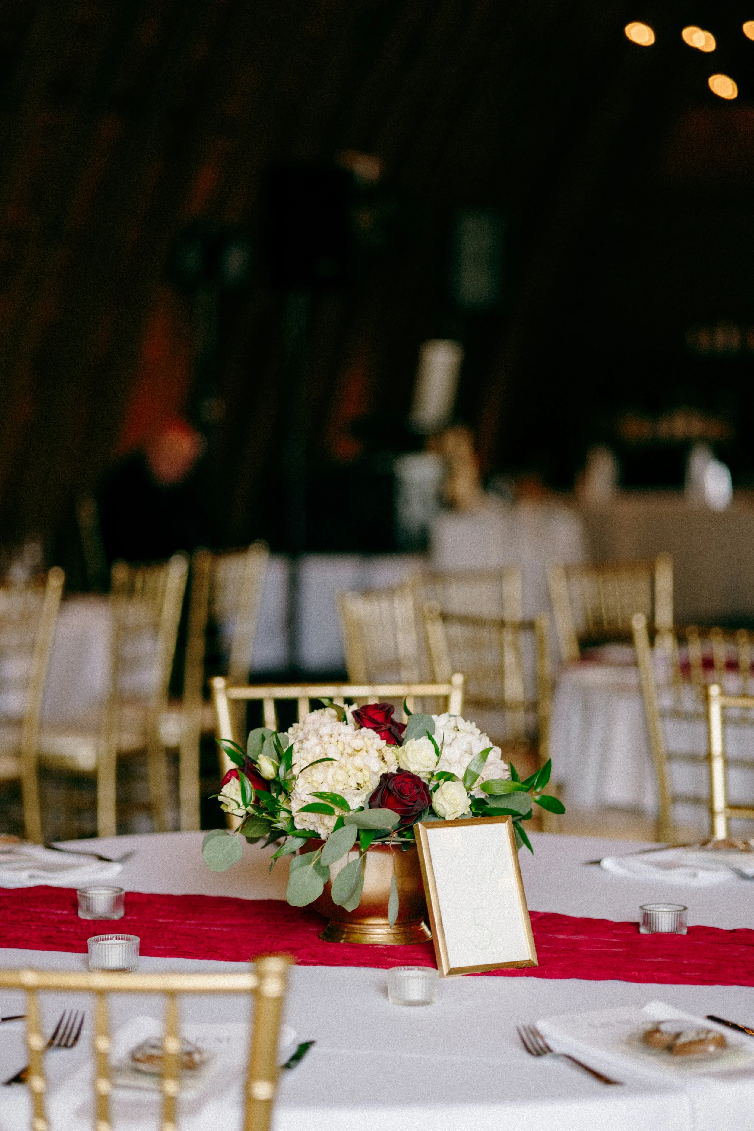 A formal banquet table decorated with a floral centerpiece of white hydrangeas, red roses, and green leaves, with a table number card in a gold frame, set in an elegant venue with gold chairs and white tablecloths.