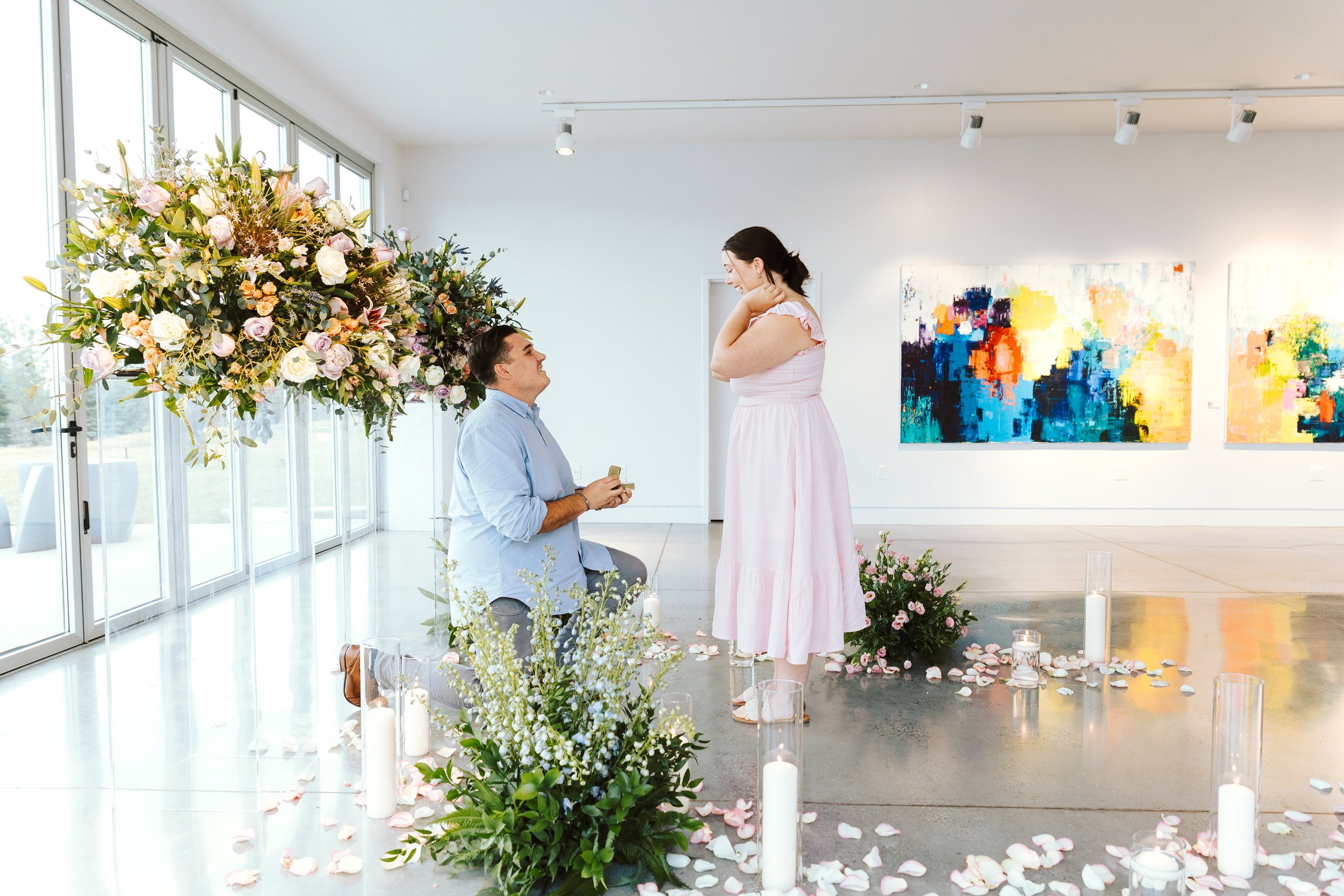 A man proposing marriage to a woman in an art gallery decorated with flowers, candles, and rose petals.
