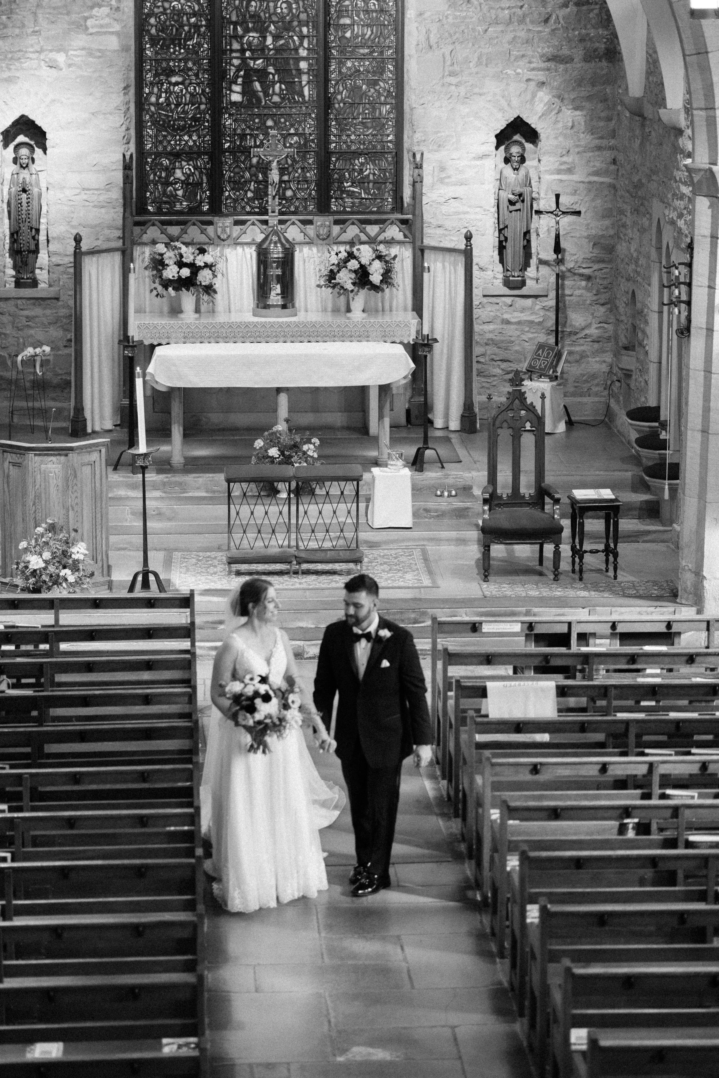 A bride and groom walking down the aisle of a church, with the altar and religious statues in the background.