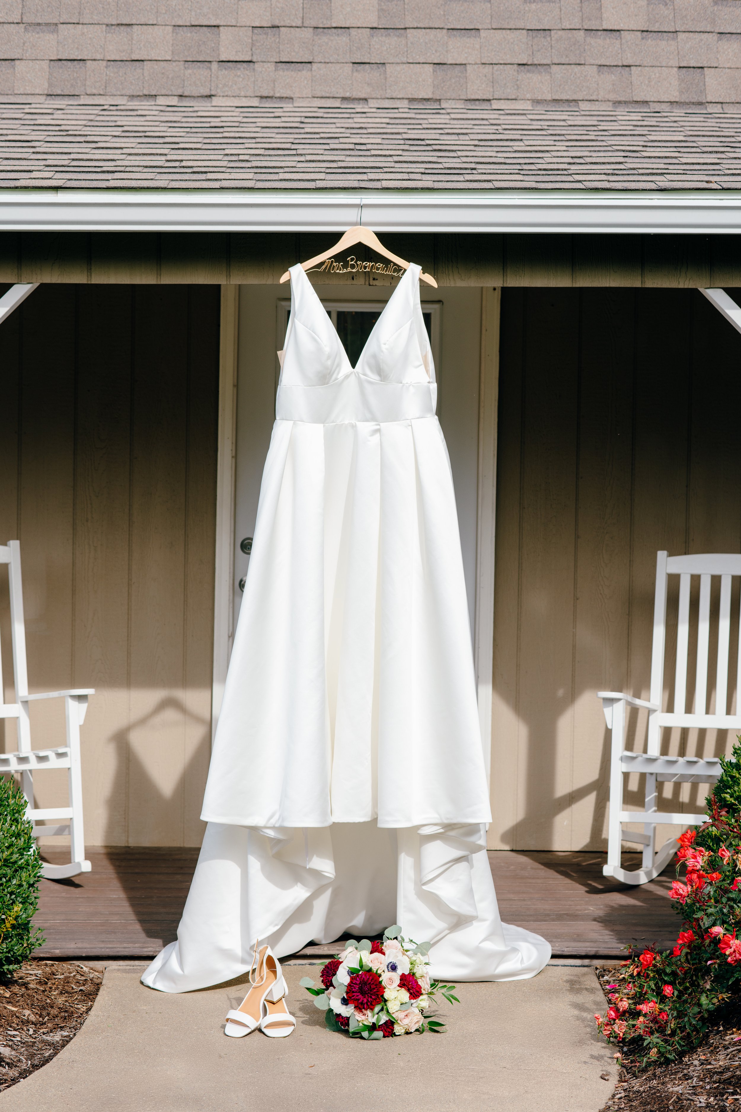 A white wedding dress hanging on a wooden hanger with a sign that reads 'Mrs. Brandau' on a porch, with a bouquet of flowers and a pair of white heels placed on the ground.