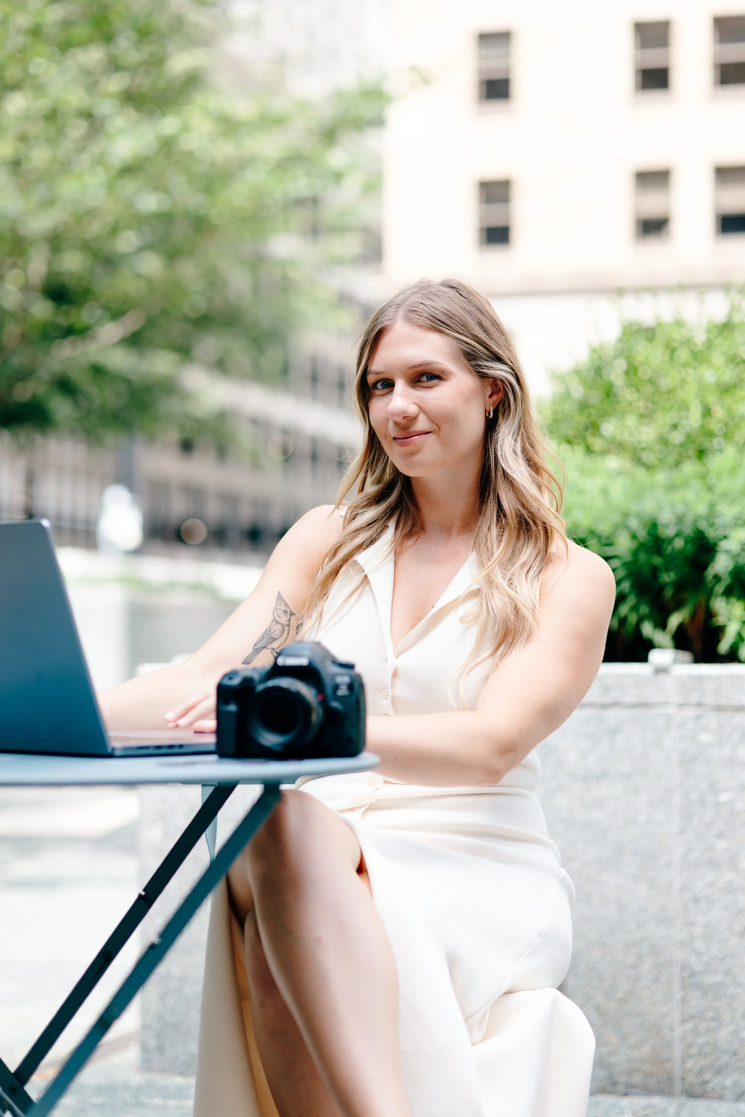 A woman in a cream sleeveless dress sitting outdoors at a small table with a laptop and camera, with green trees and city buildings in the background.