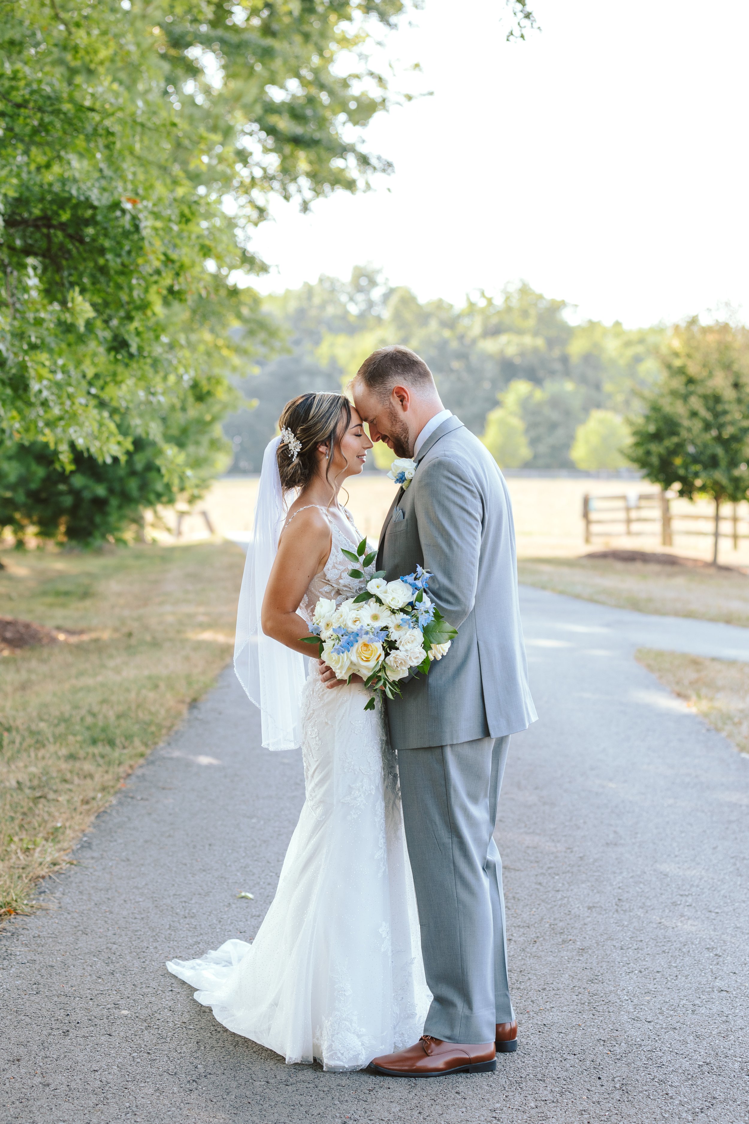 A bride and groom stand close together on a paved path in a park, holding a bouquet of white and blue flowers with greenery, in a loving embrace, touching foreheads with eyes closed, during their wedding.