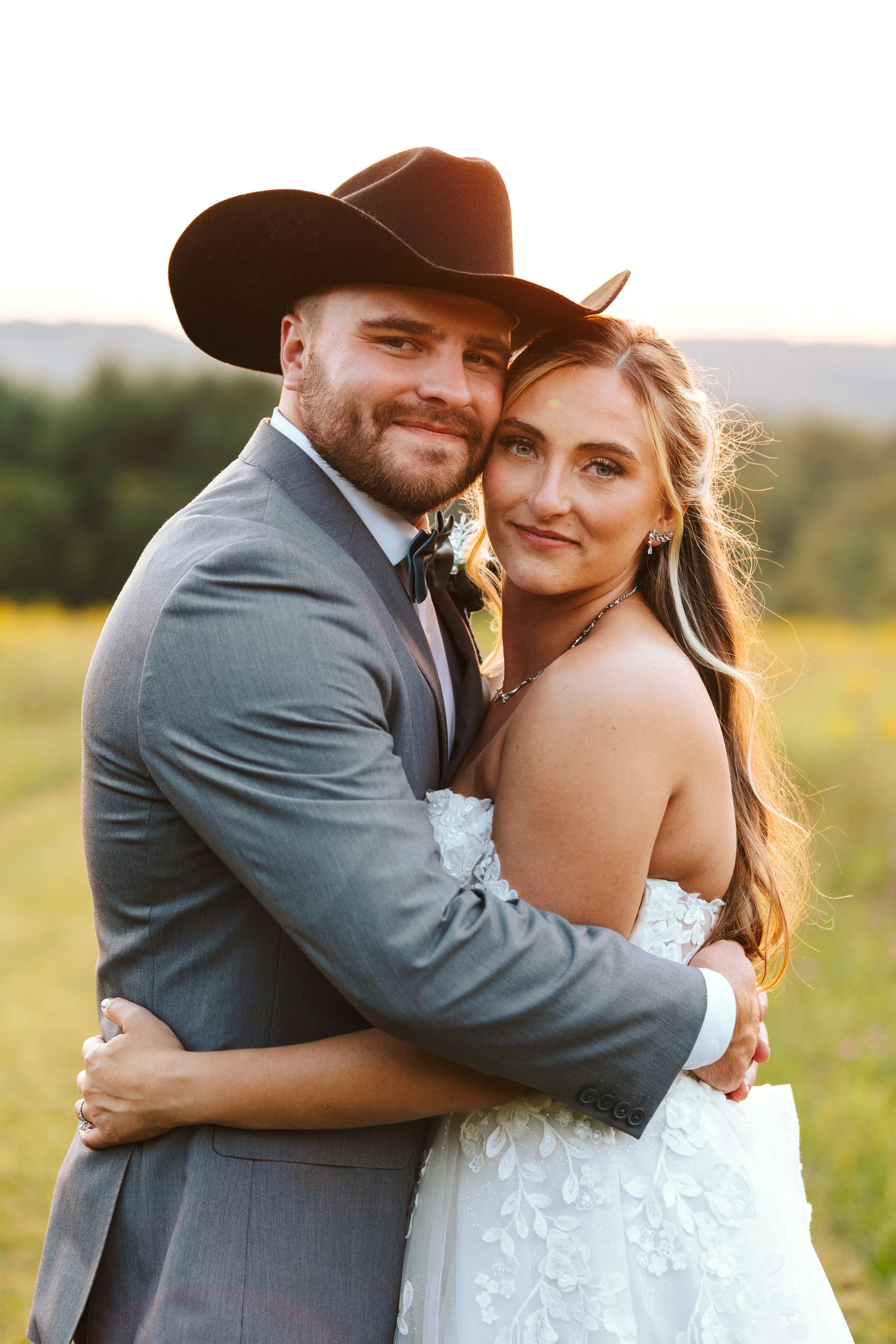 A newlywed couple is embracing outdoors during sunset, with the man wearing a gray suit and cowboy hat, and the woman in a white lace wedding dress, both smiling at the camera.