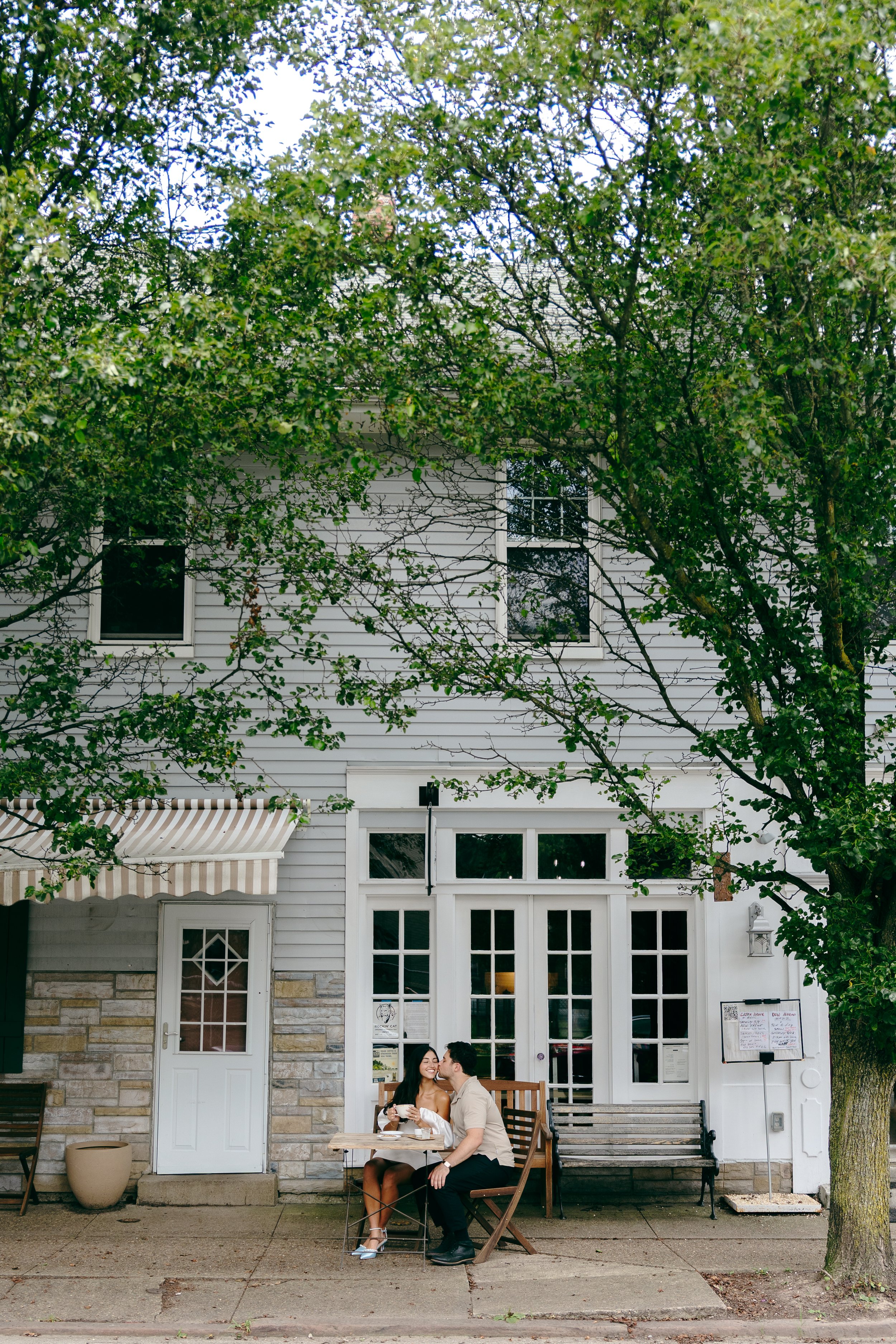 A couple sitting at an outdoor table in front of a white building with large windows, surrounded by trees.