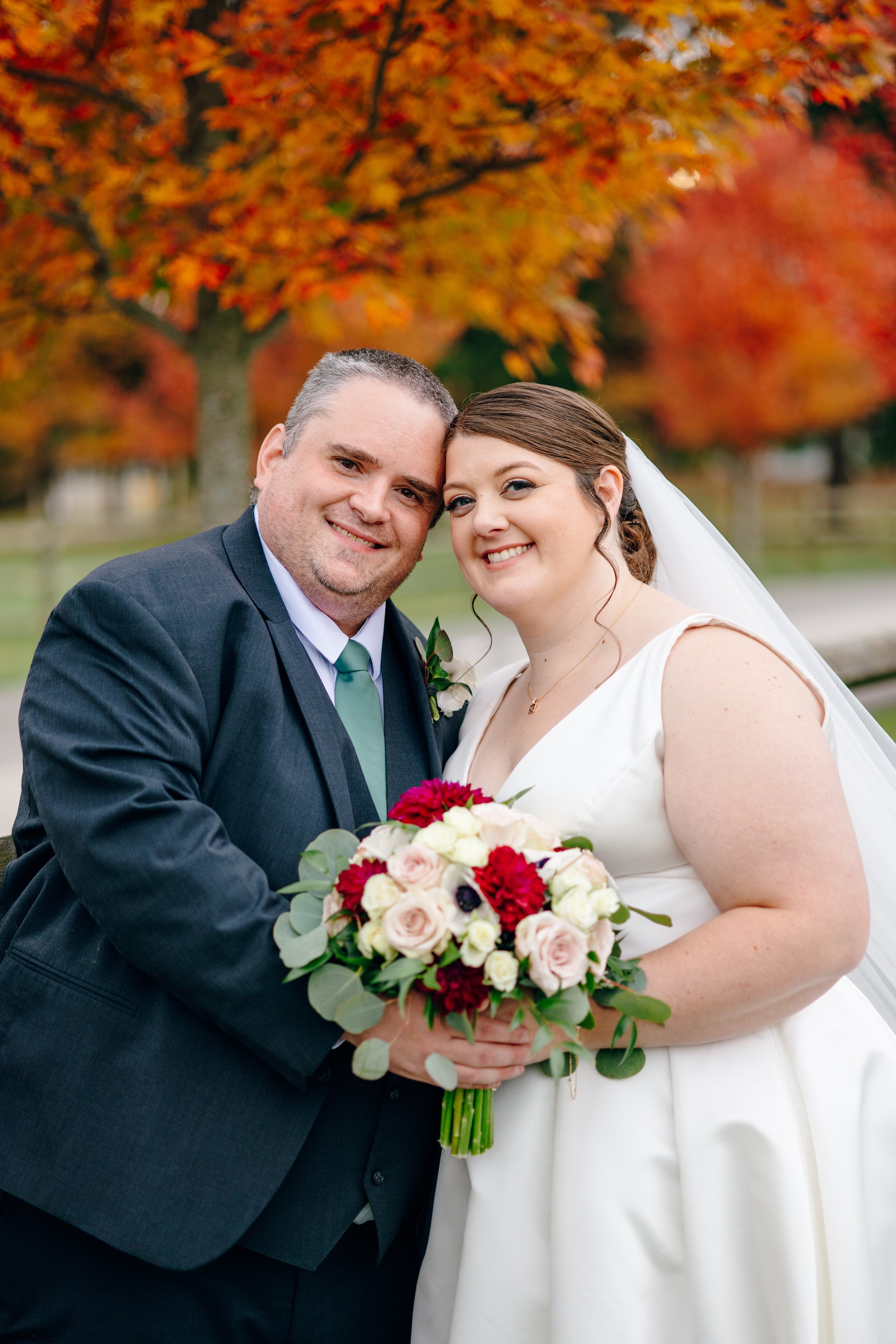 A newlywed couple smiling, holding a bouquet of flowers, standing outdoors with autumn leaves on trees in the background.