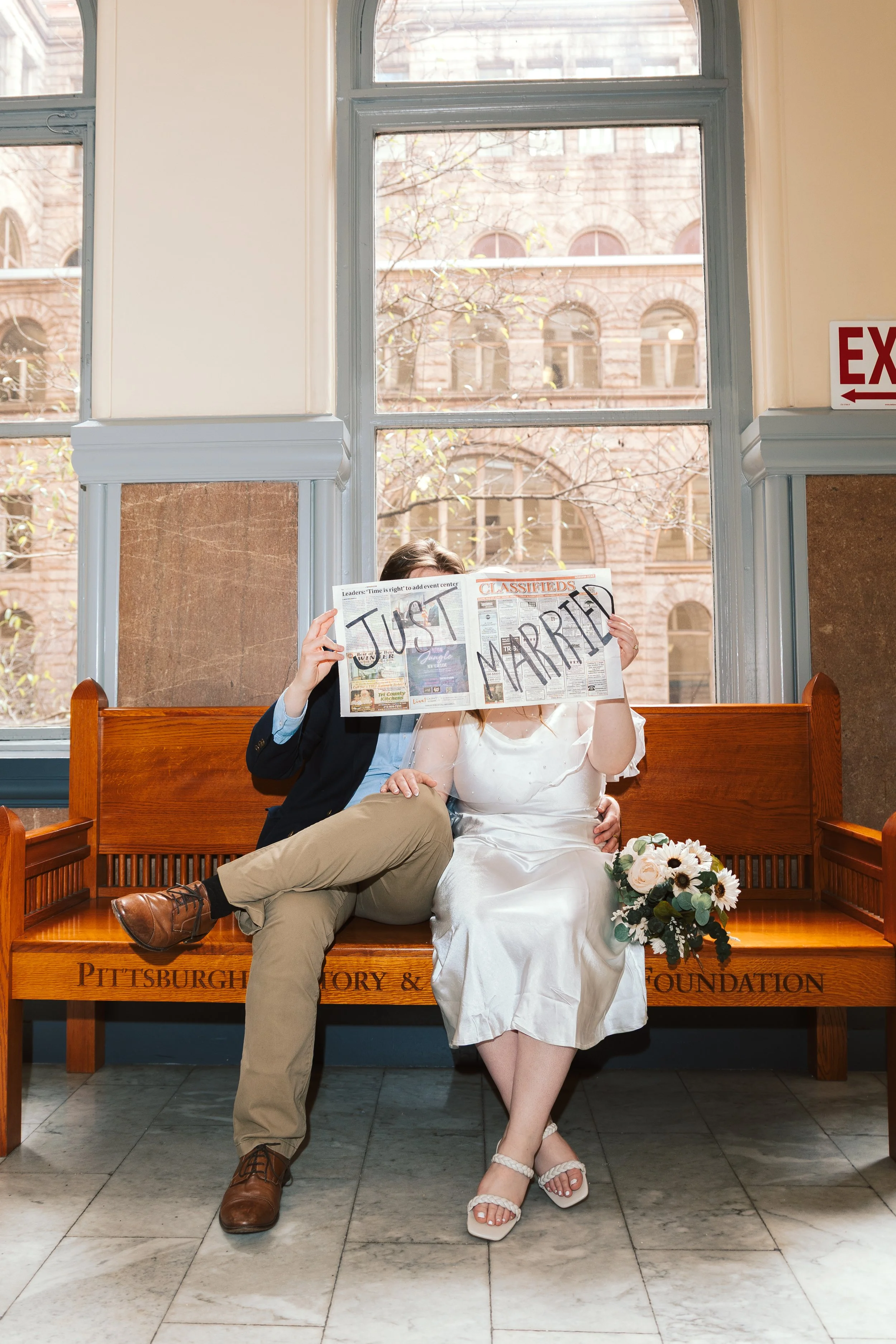 A couple sitting on a wooden bench, the person on the left holding a newspaper in front of their face that reads "JUST MARRIED". The woman on the right is dressed in a wedding gown, holding a bouquet of flowers.