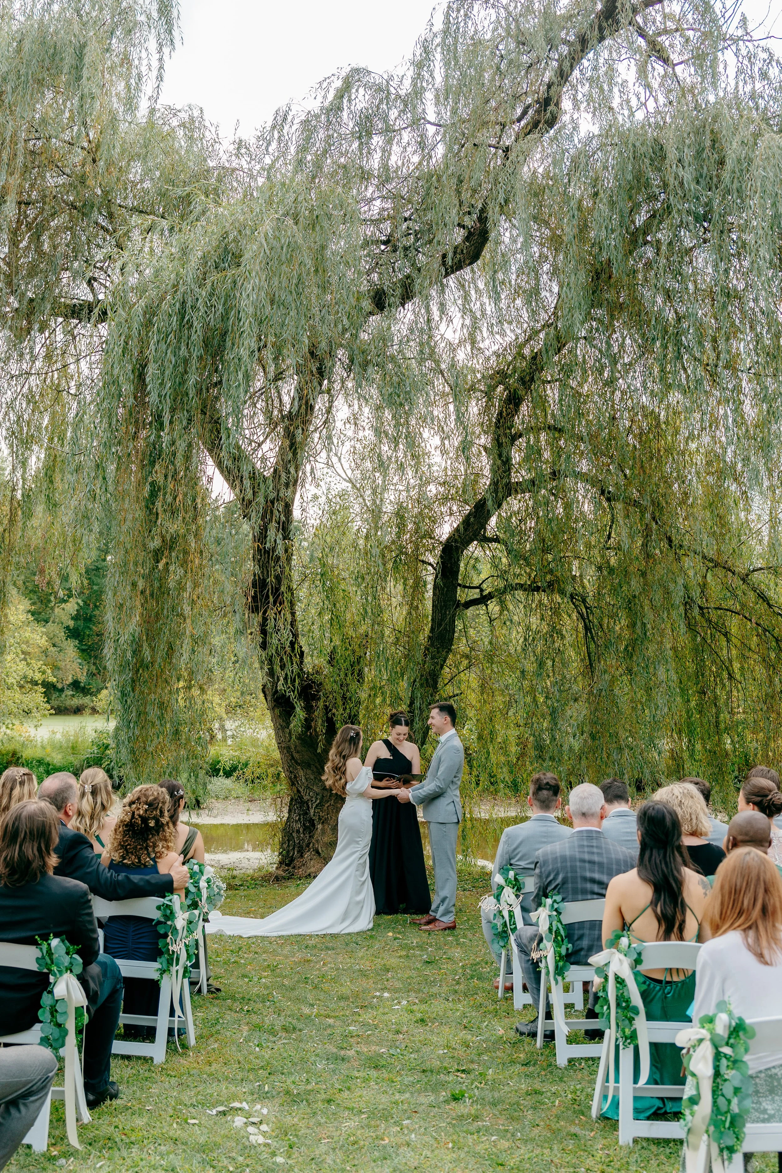 A wedding ceremony outdoors under a large tree with a bride and groom exchanging vows, surrounded by seated guests decorated with greenery and ribbons.