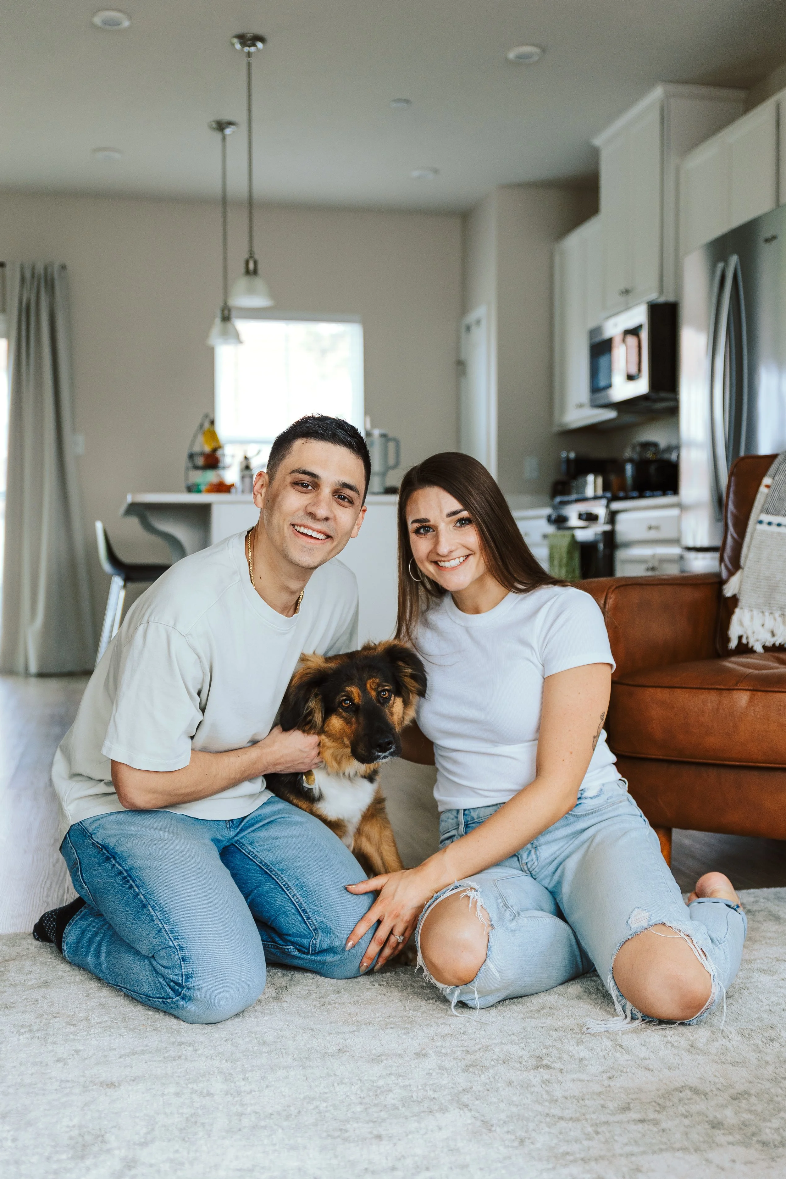 A happy couple with a dog sitting on the floor of their living room, smiling at the camera.