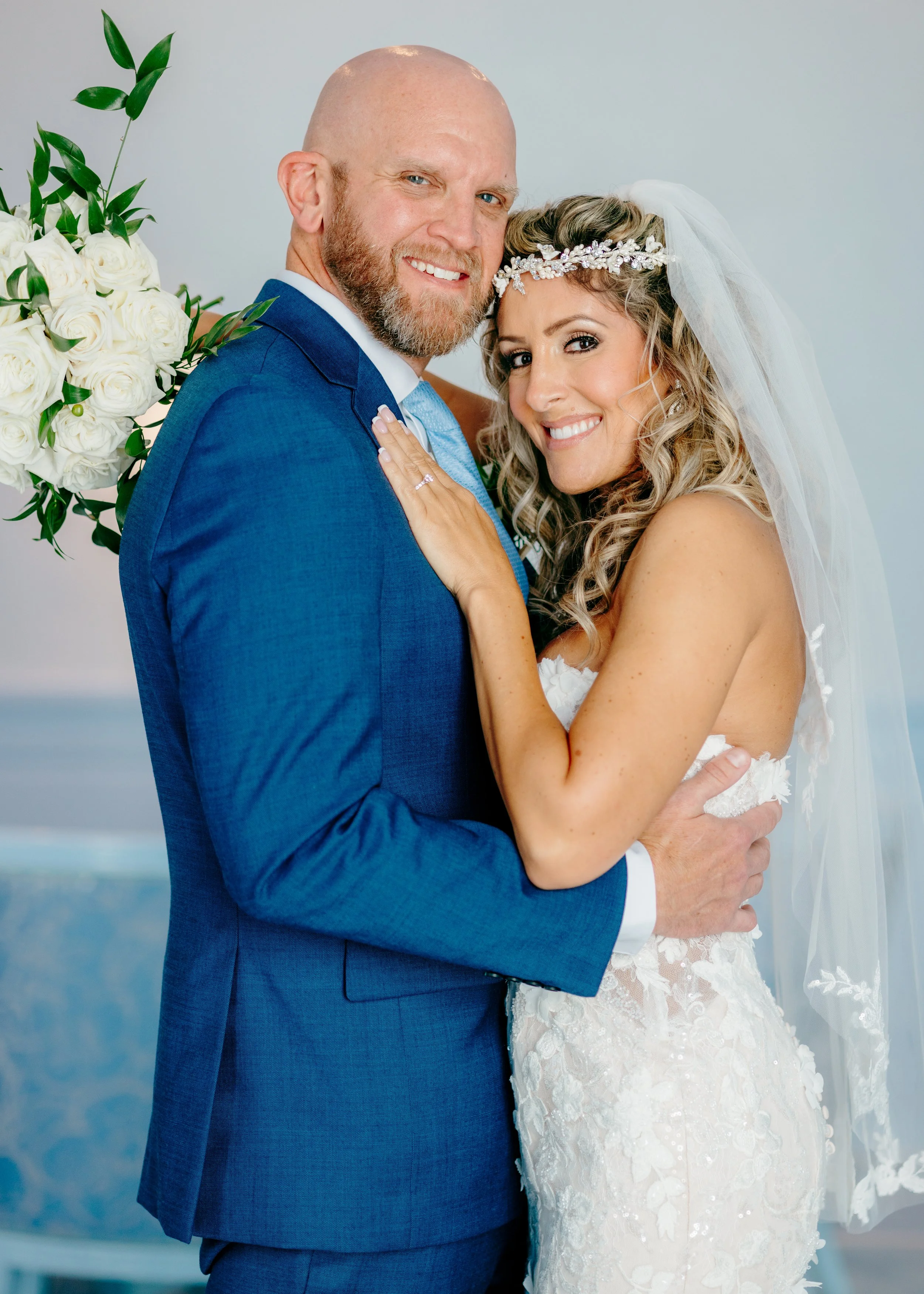 A bride and groom smiling and embracing each other, with the bride wearing a white wedding dress and veil, and the groom in a blue suit, during their wedding celebration.