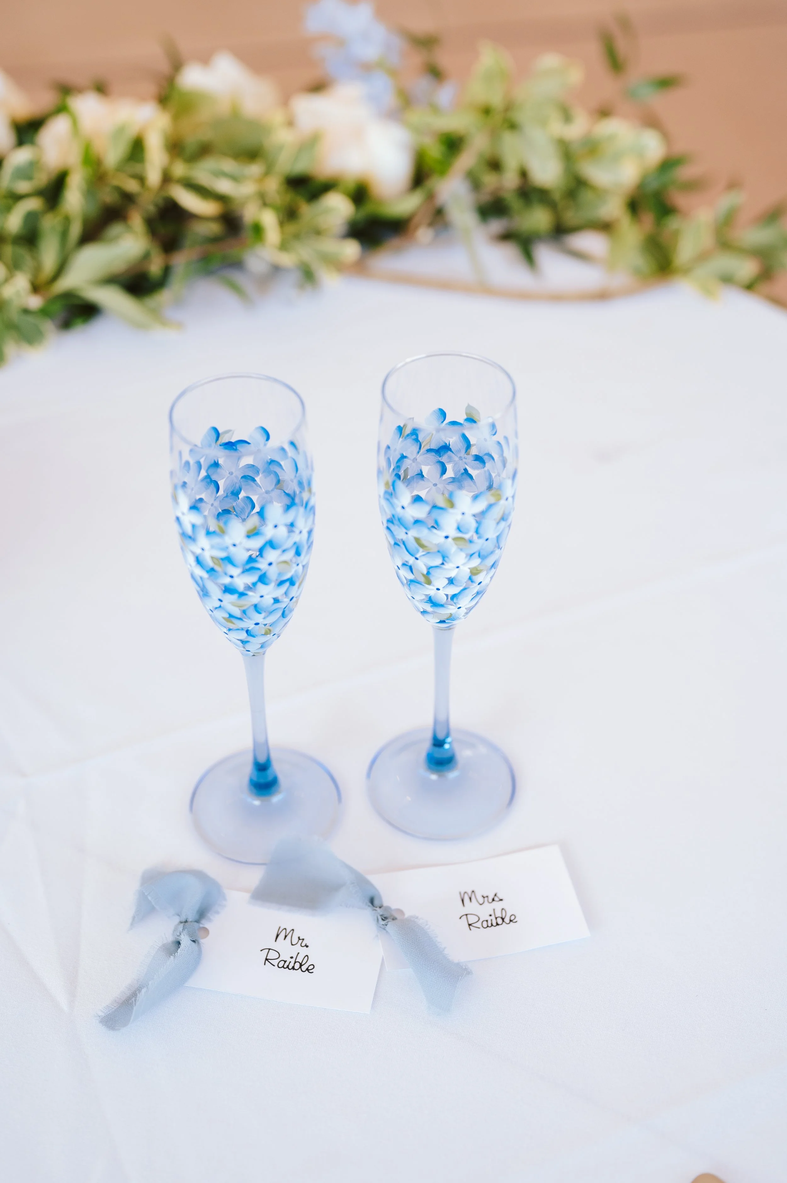 Two champagne flutes filled with small blue and white flower petal decorations, placed on a white table with name tags labeled 'Mrs. Raible' tied with light gray ribbons.