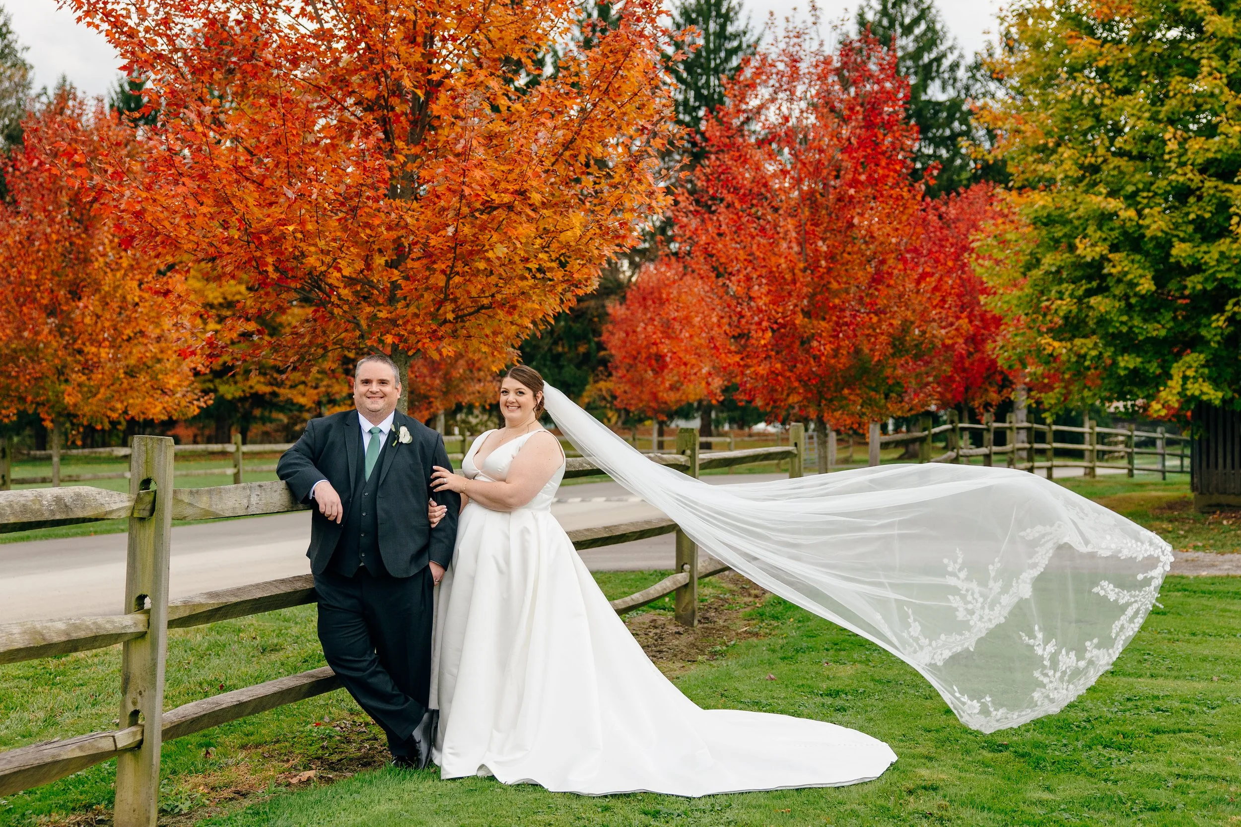 A newlywed couple stands near a wooden fence in front of vibrant autumn trees, with the bride wearing a white gown and veil, and the groom in a dark suit with a white boutonniere.