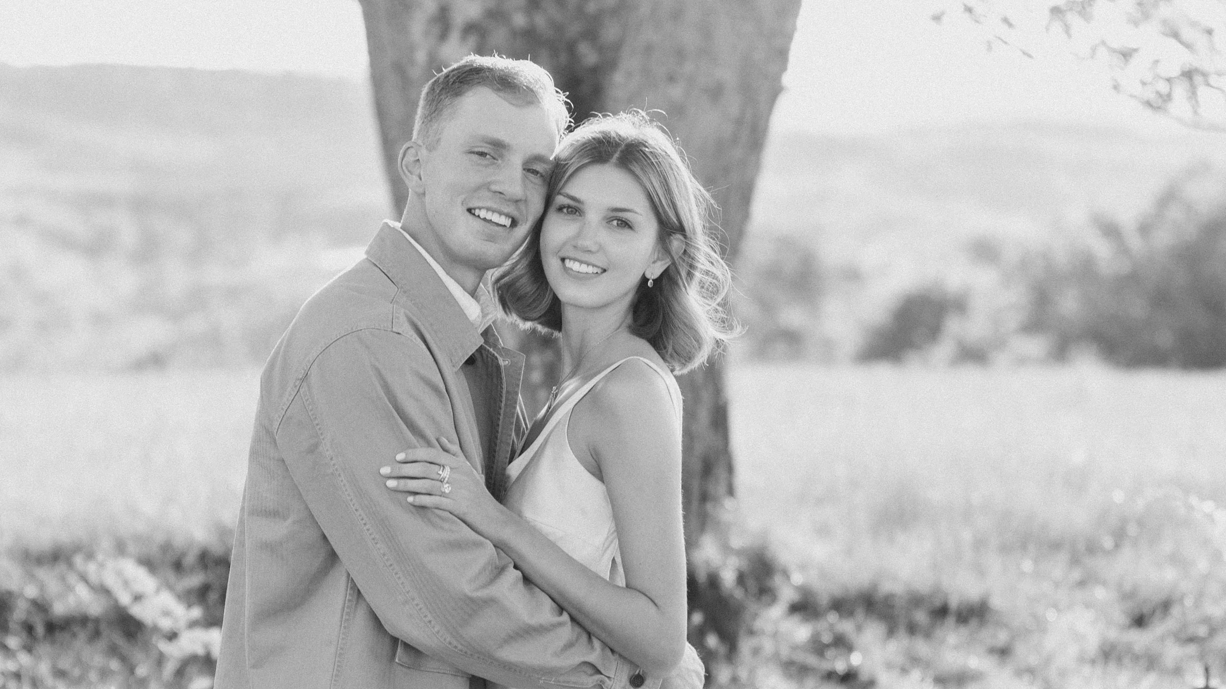A smiling couple standing outdoors near a tree in a grassy field.