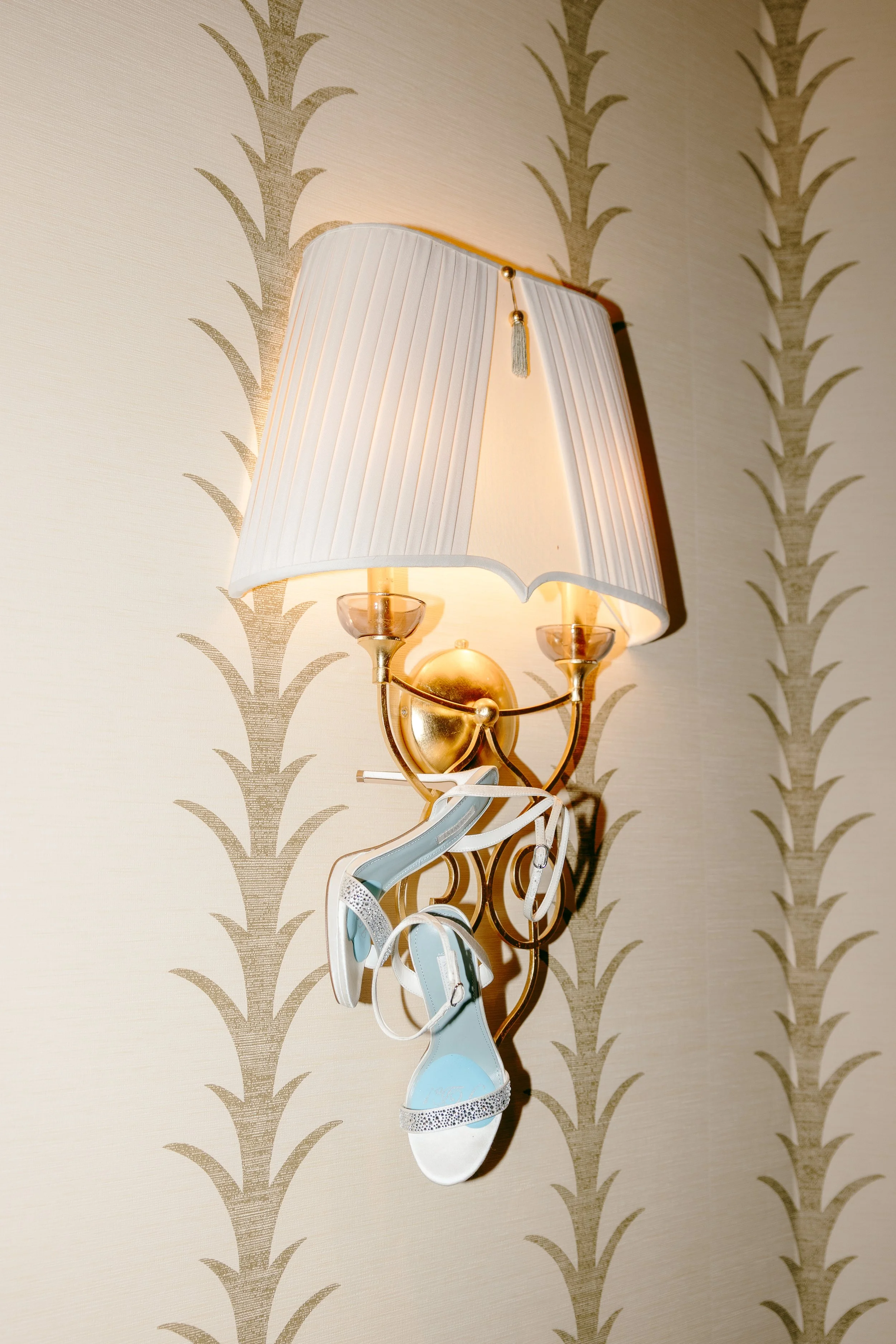A wall-mounted gold-colored lamp with a white lampshade and a tassel ornament. A pair of white and light blue high-heeled sandals with rhinestone details dangle from the lamp.