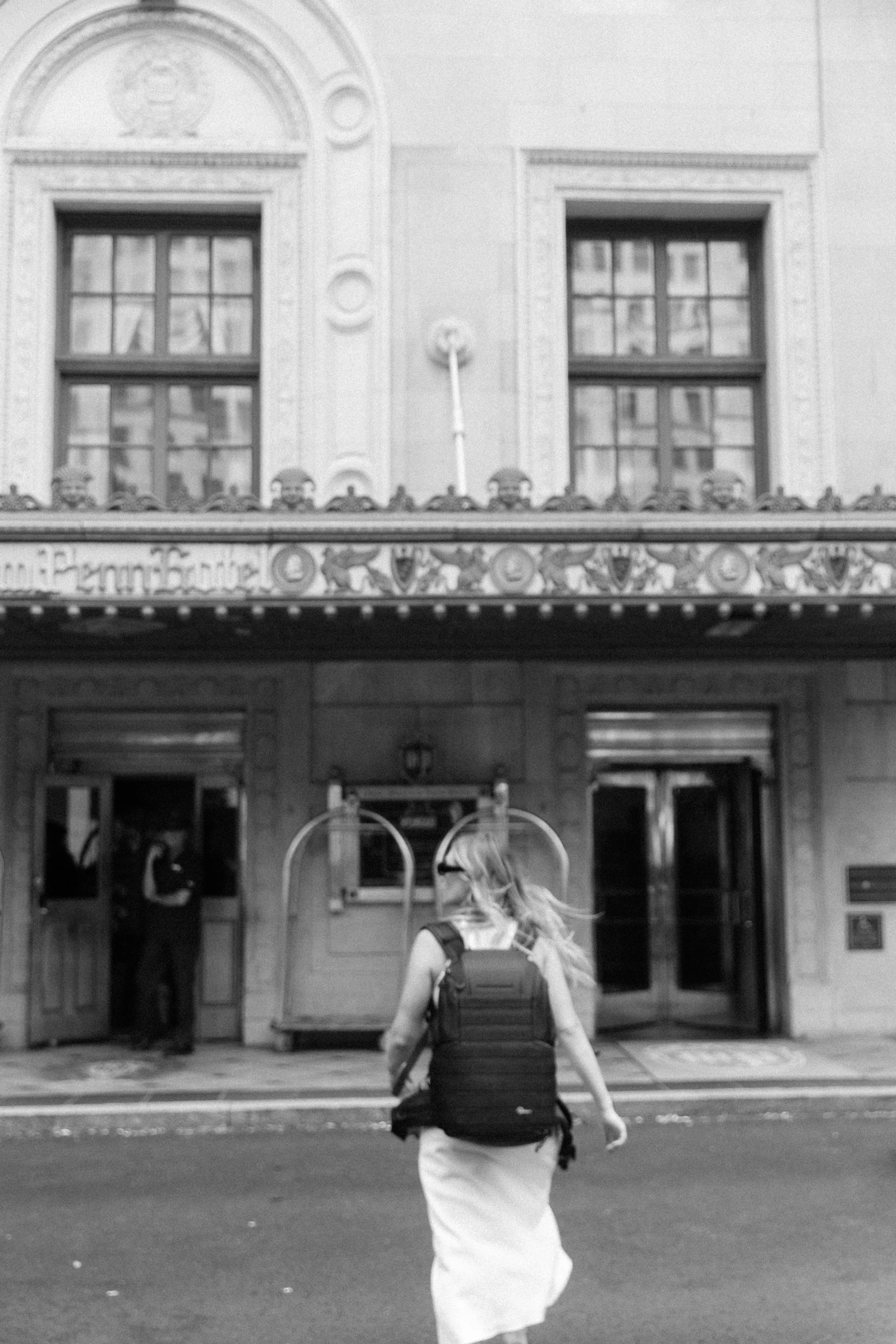 A woman with long hair and a backpack walking towards a building with ornate architecture in black and white.