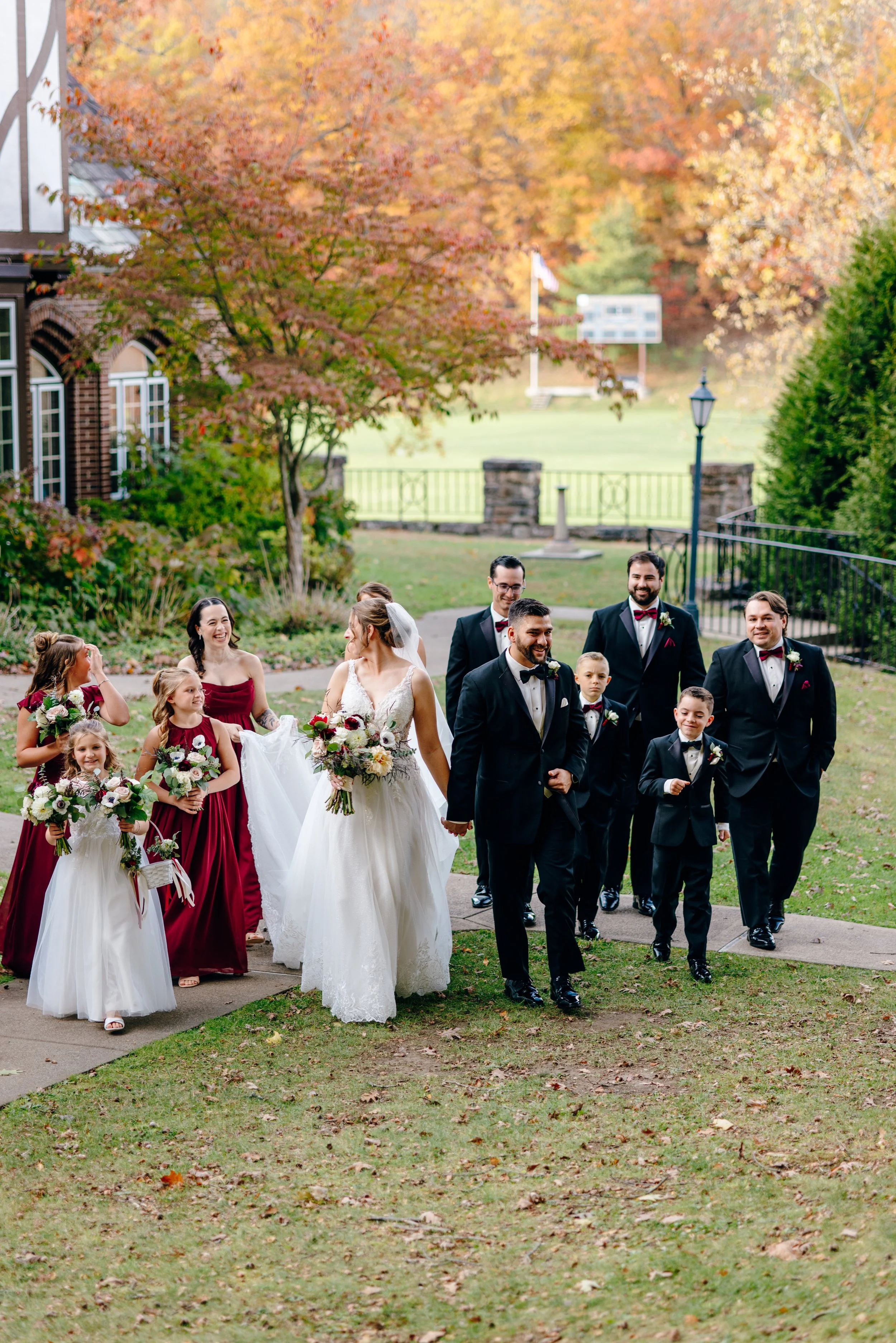 A wedding party walking outside on a fall day with colorful autumn trees in the background. The bride is in a white wedding gown holding a bouquet, surrounded by bridesmaids in burgundy dresses and children also in formal attire. Groomsmen in black suits and bowties are part of the group.