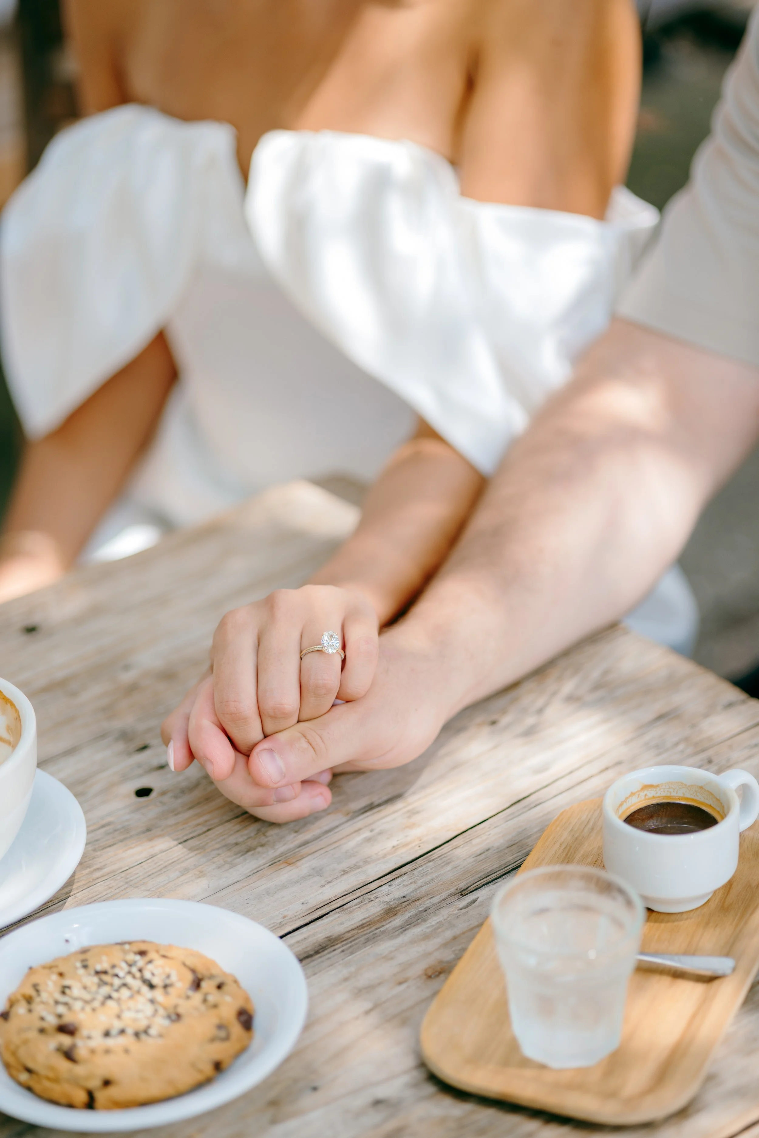 Person wearing a white dress with puffed sleeves, showing an engagement ring while holding hands with another person at a wooden table with coffee, a cookie, and water.