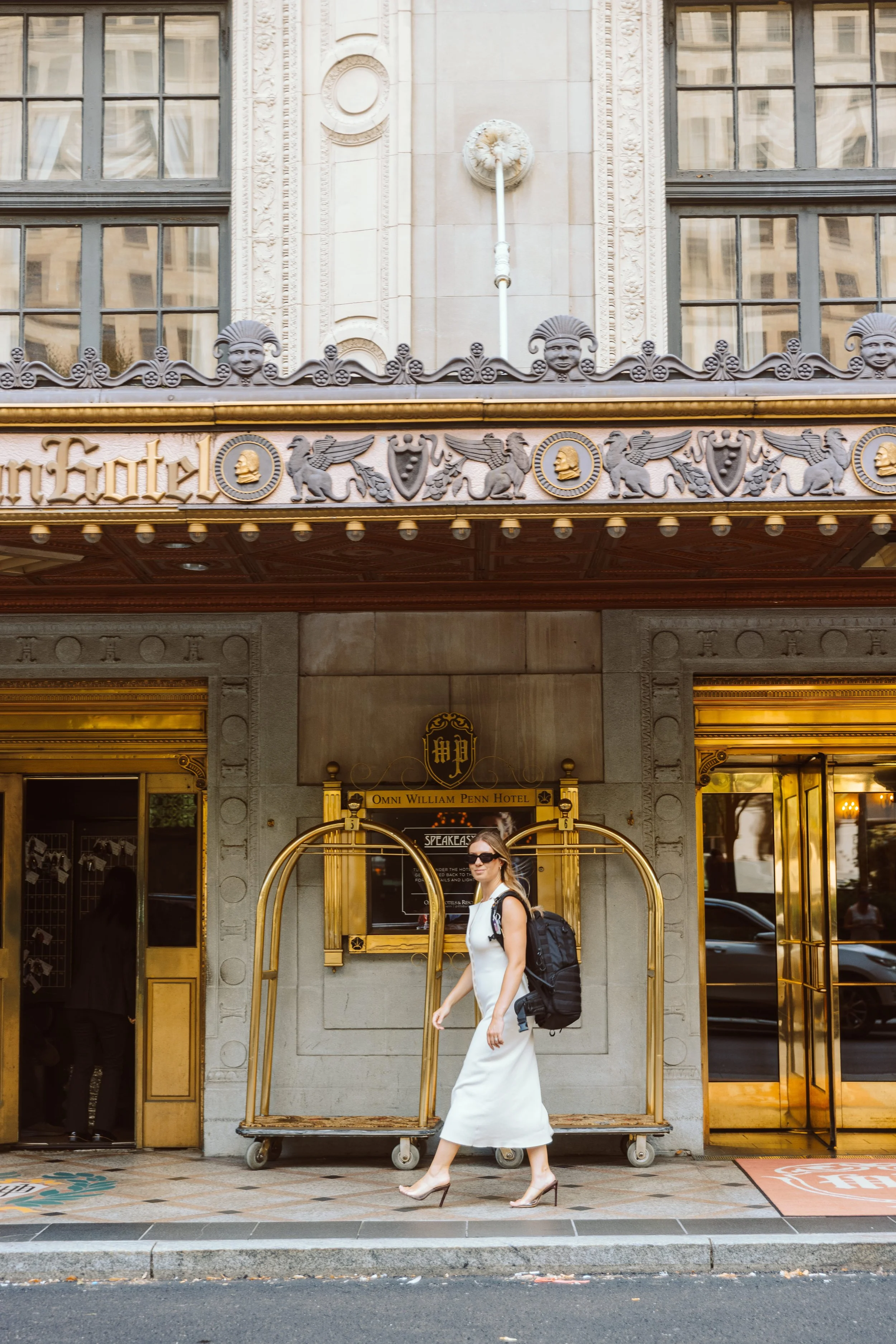 A woman in a white dress and high heels walking past the entrance of the Omni William Penn Hotel, which has ornate gold and gray architectural details and a gold luggage cart outside.