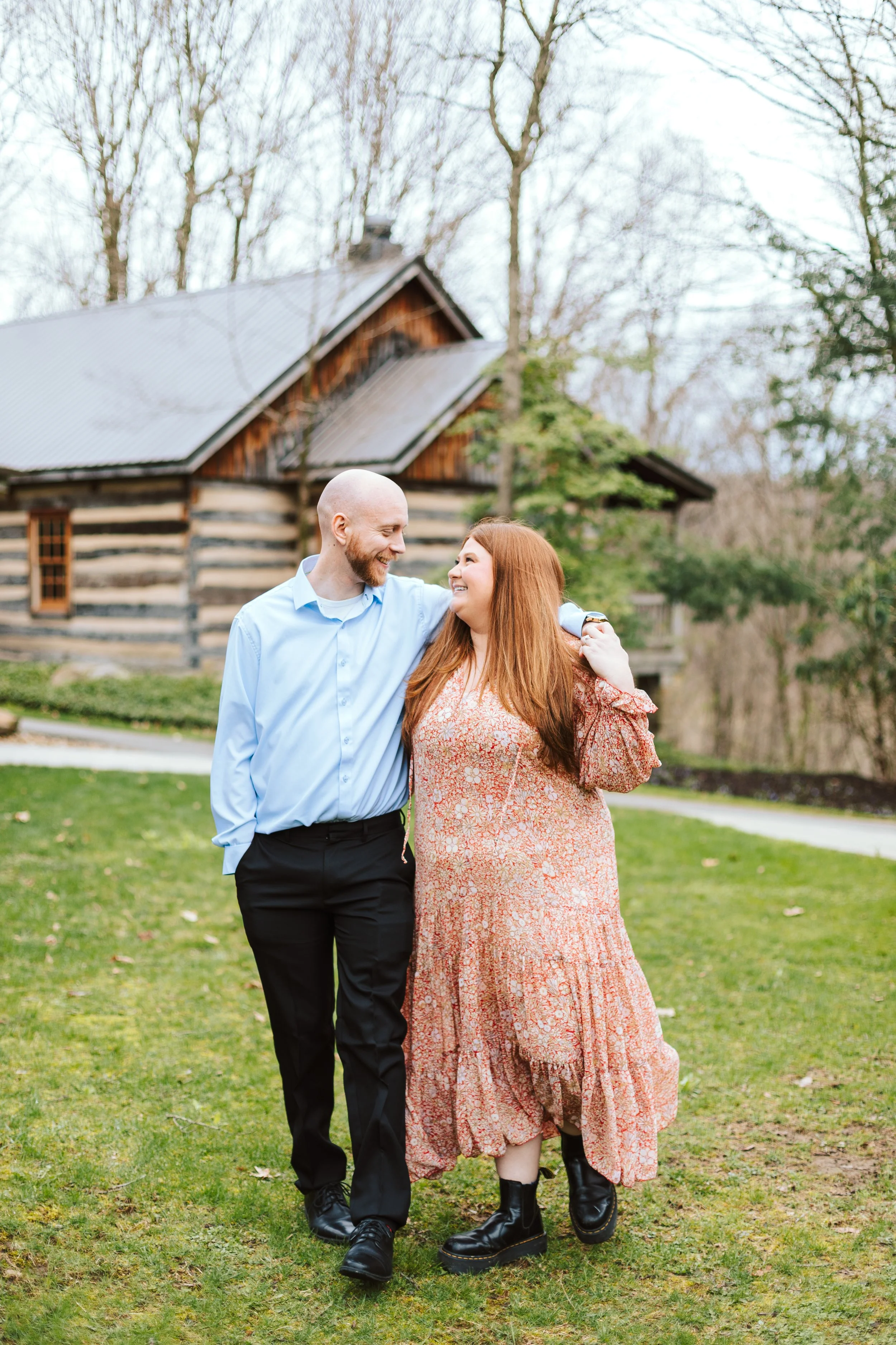 A couple walking on grass near a rustic cabin, smiling at each other.
