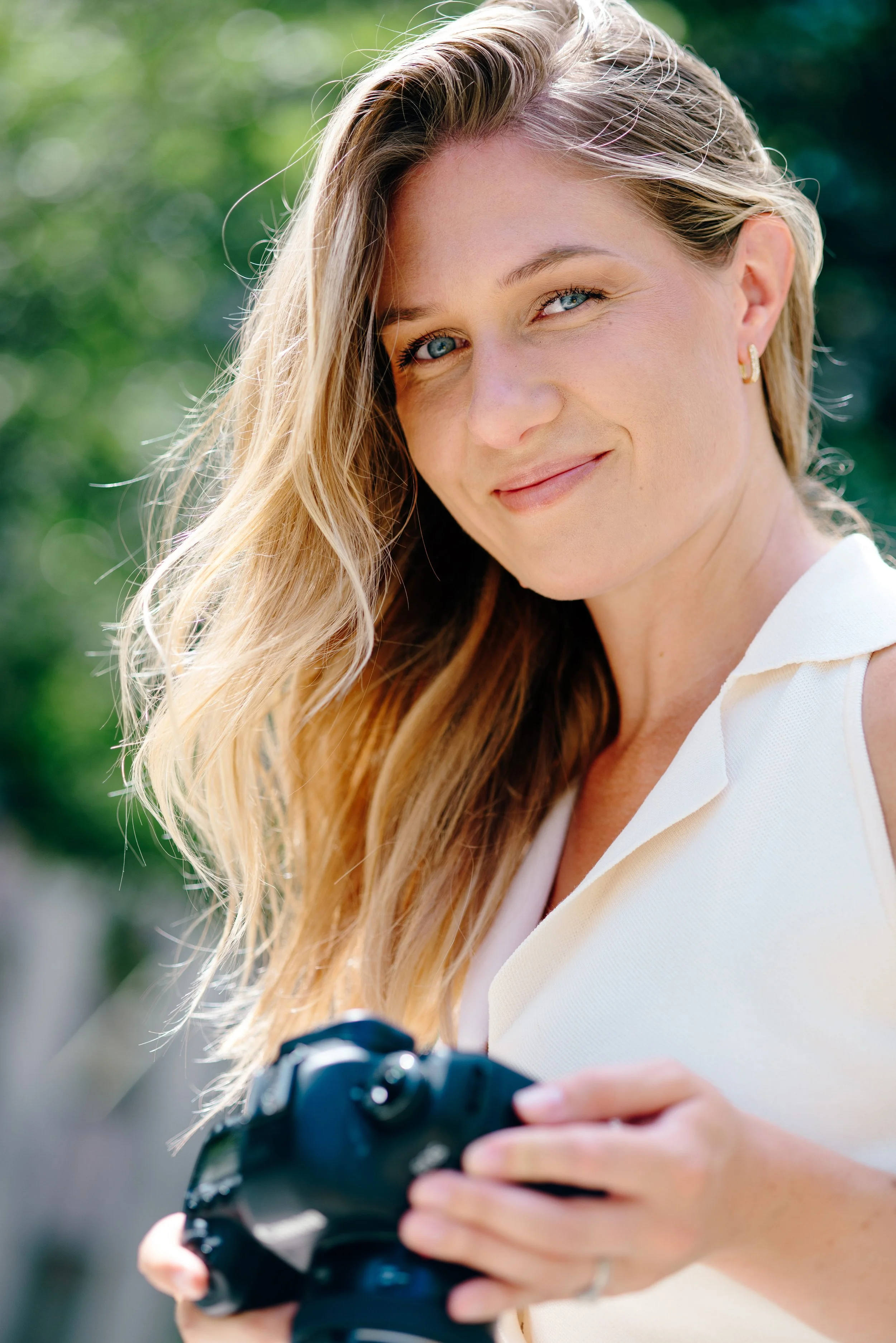 A young woman with long, wavy blonde hair smiling and holding a camera outdoors with a blurred green background.