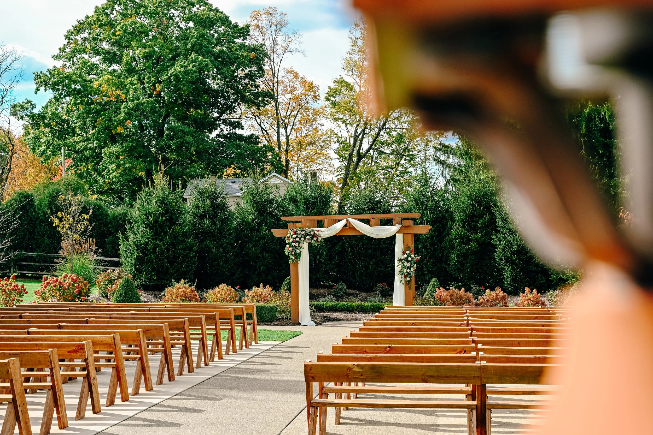 Outdoor wedding ceremony setup with wooden chairs arranged on either side of a central aisle, a decorated wooden arch with white fabric and floral arrangements, surrounded by lush green trees and plants, with partly cloudy sky in the background.