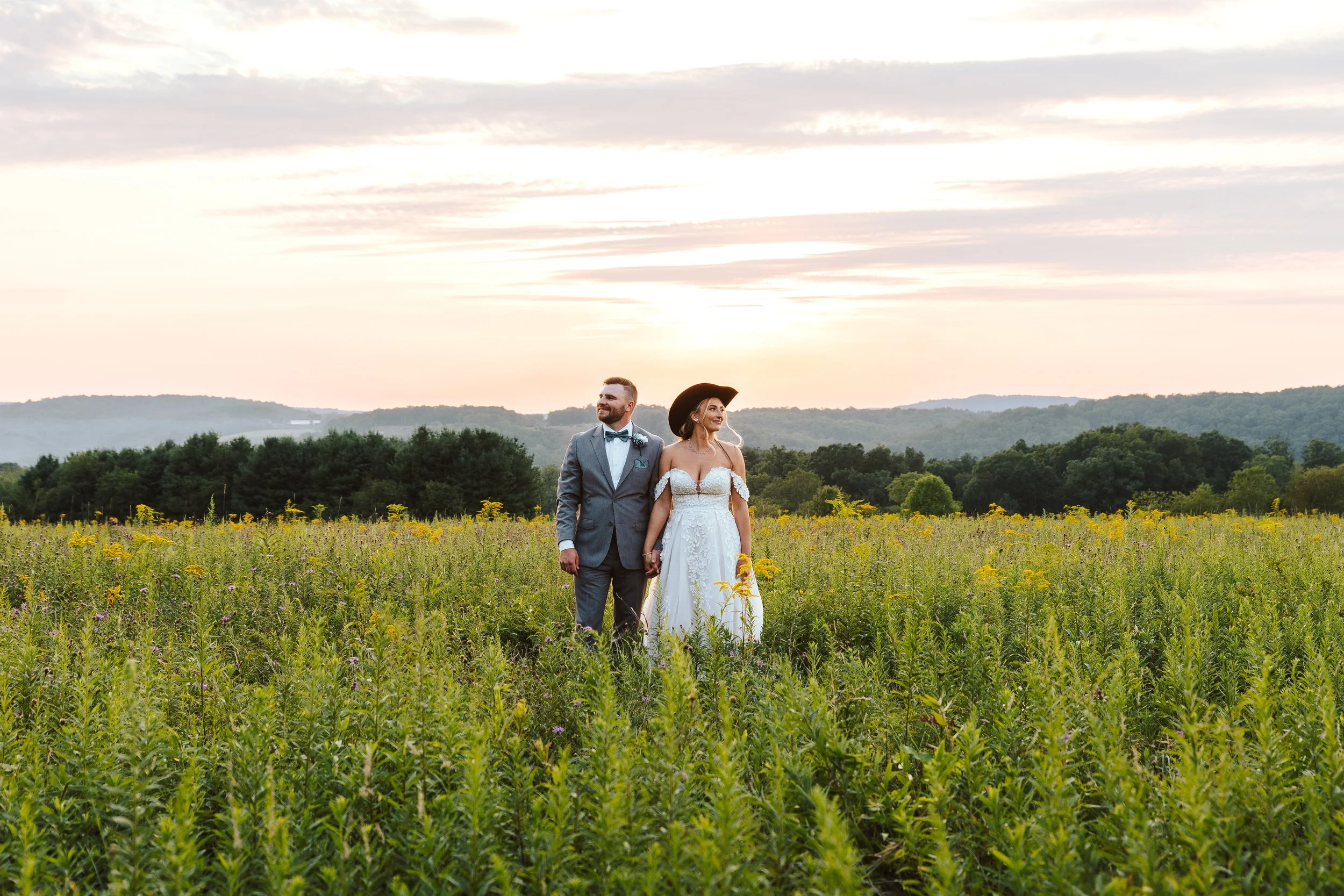 A couple in wedding attire holding hands in a green field at sunset, with mountains and trees in the background.