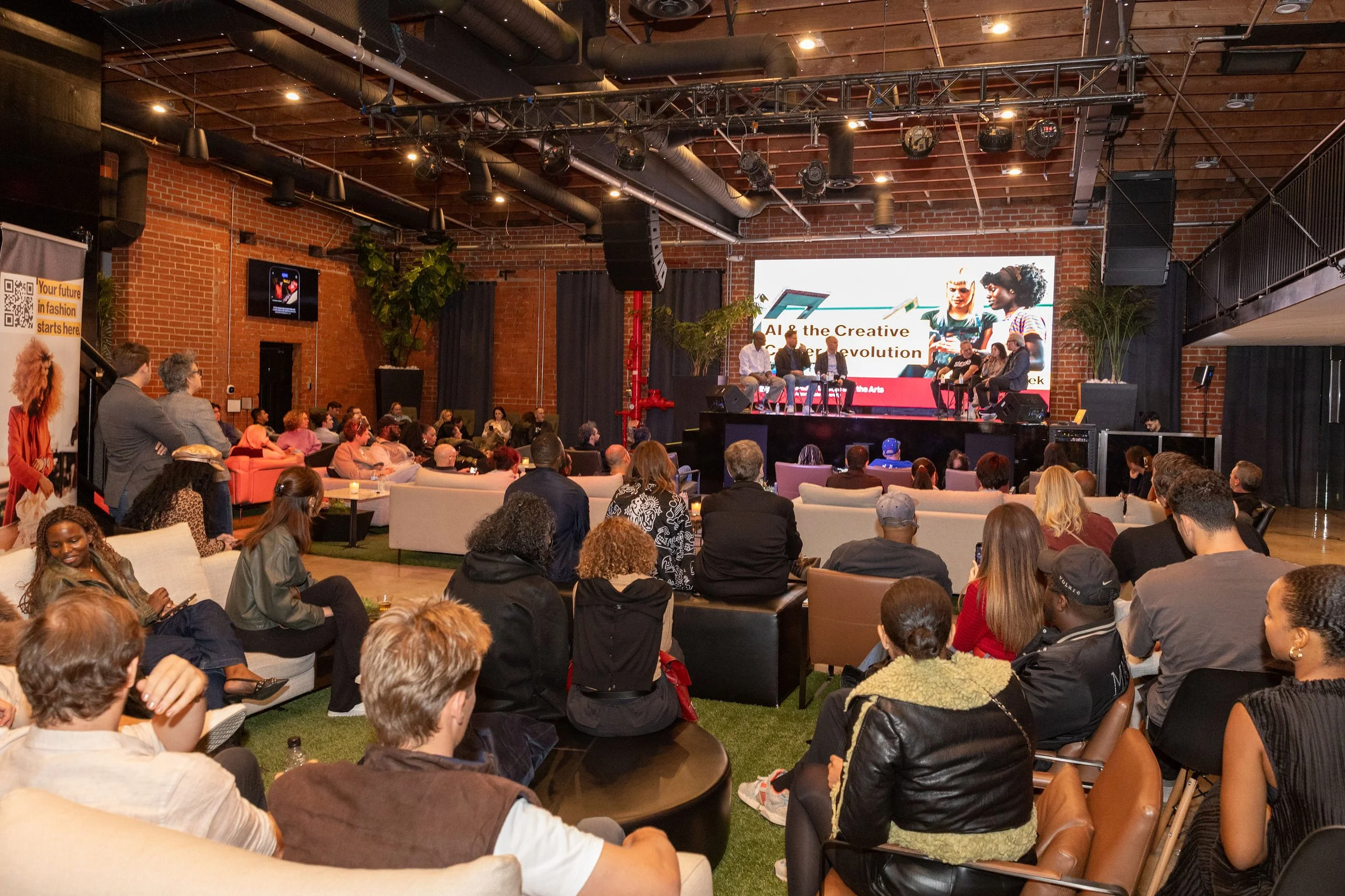 Audience watching a panel discussion on stage in a large, brick-walled venue with a digital screen displaying the event title, 'AI & the Creative Revolution,' as speakers participate in a panel discussion.