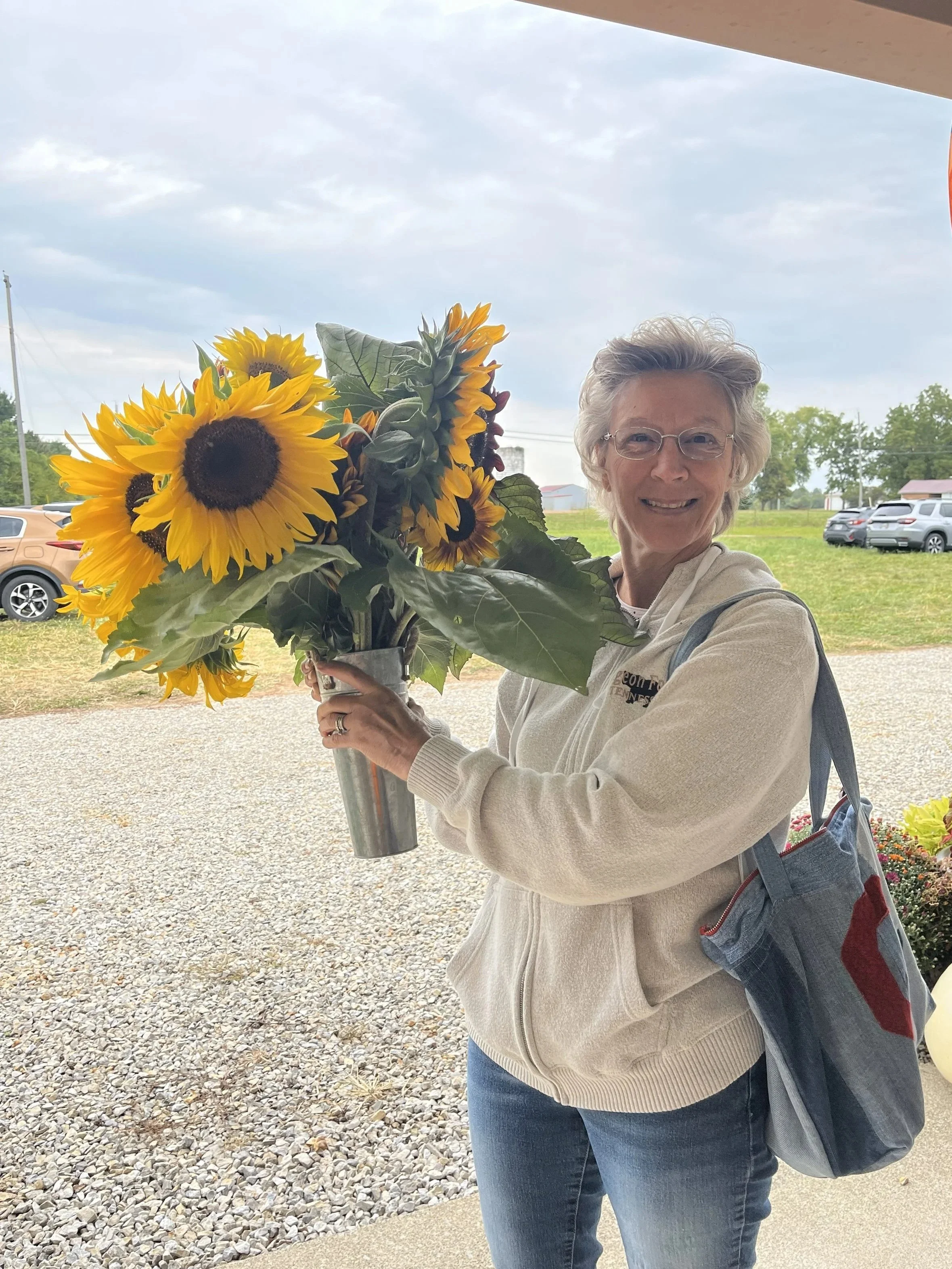 A smiling woman holding a large bouquet of sunflowers outdoors, with a parking lot and green landscape in the background.