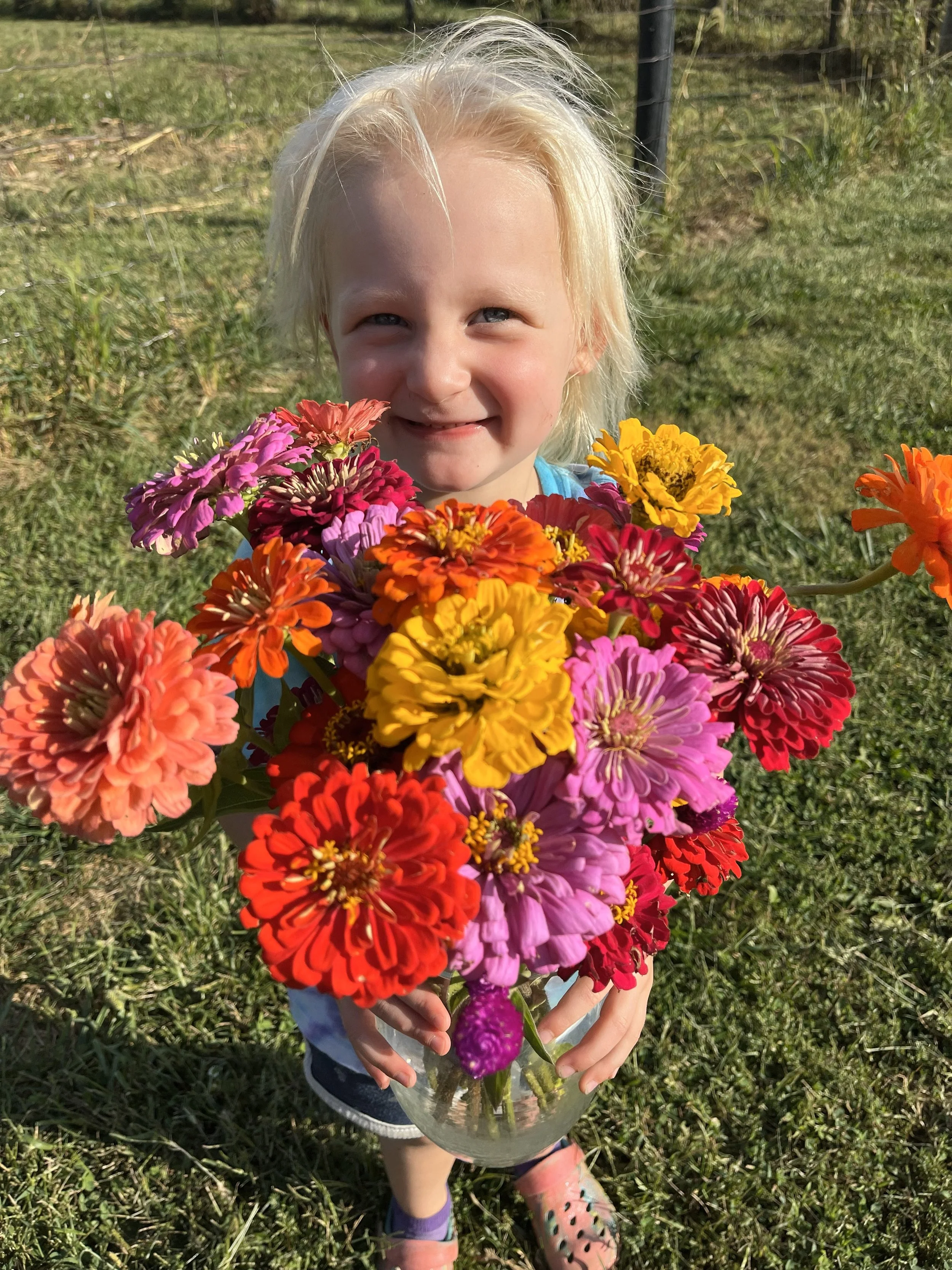 A young girl with blonde hair holding a colorful bouquet of flowers in a clear glass vase outdoors on grass.