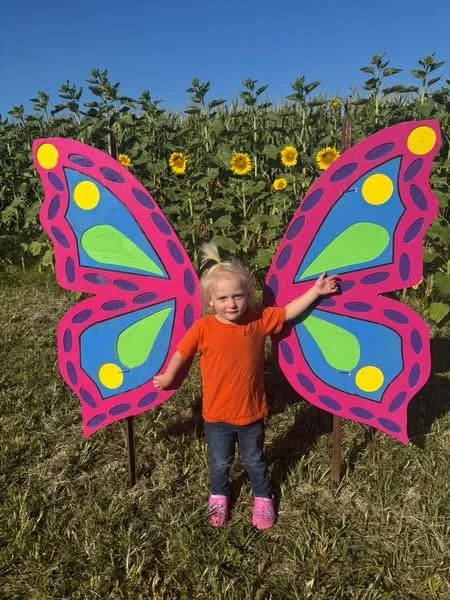 A young girl standing in front of a colorful butterfly-shaped sign with large pink and blue wings, pointing at the sky with a sunflower field and blue sky in the background.