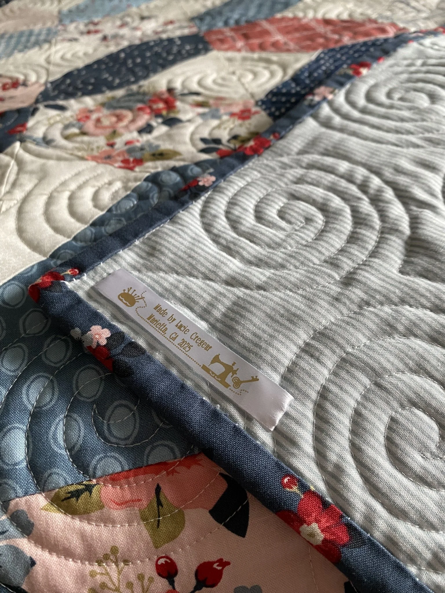 Close-up of a quilt with floral and swirl patterns, featuring a label that says it was made by Lucie Cregeur in Marietta, Georgia, with hand-stitched binding along the edge.