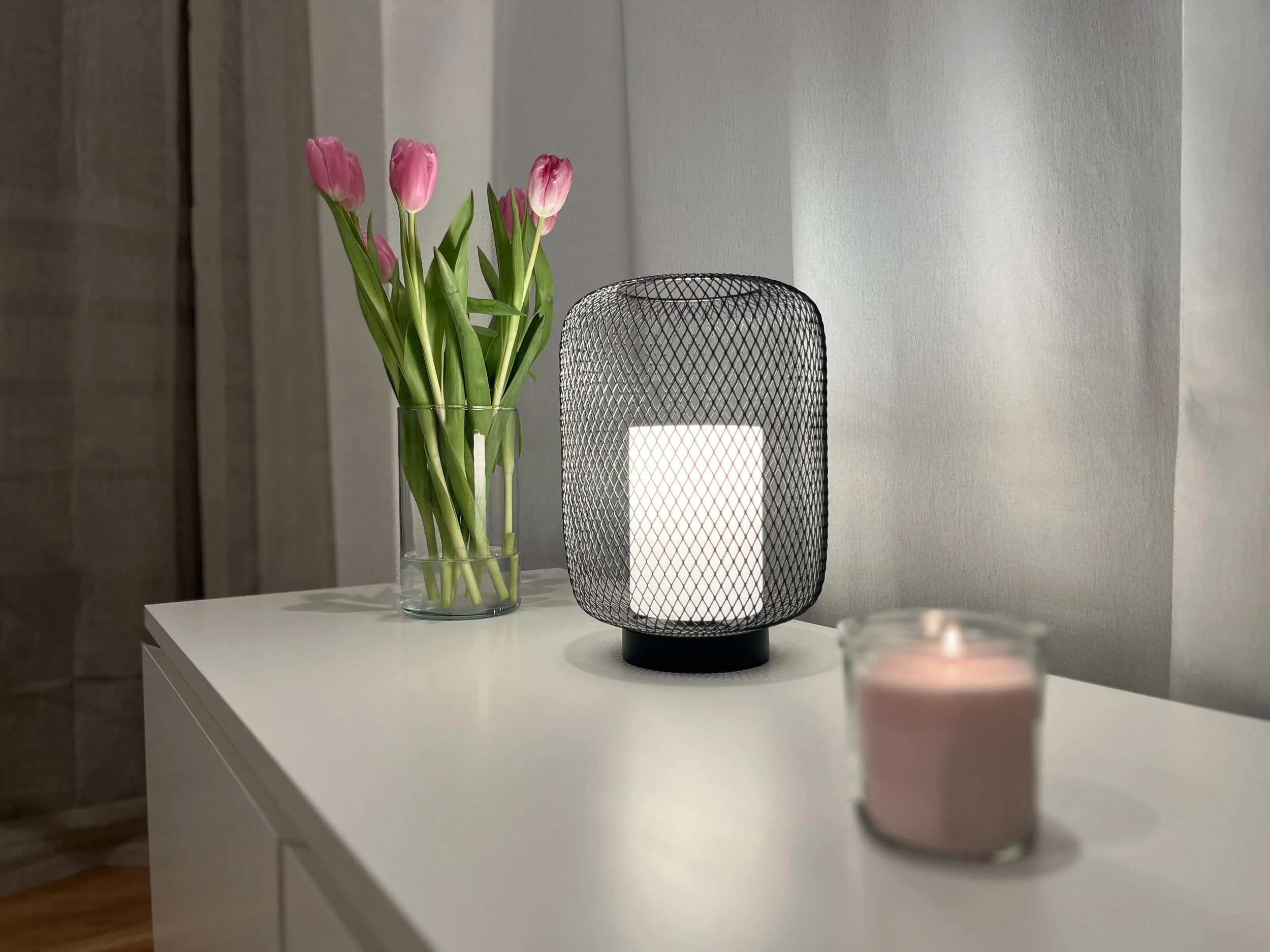 Vase mit pinken Tulpen, eine Tischlampe mit Gitter-Design und ein Kerzenlicht auf einem weißen Tisch in einem Raum mit neutralen Wänden.