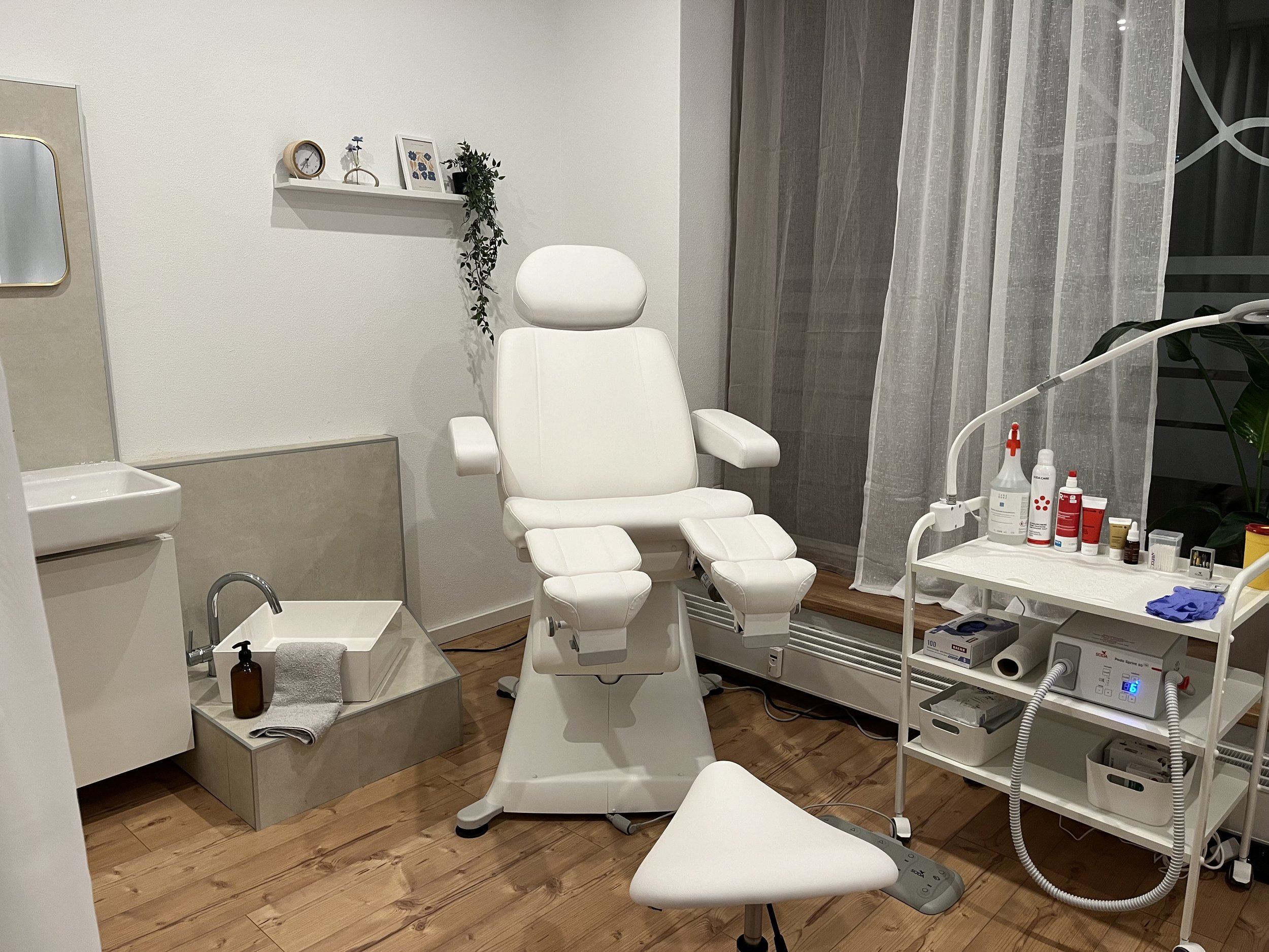 Ein Behandlungsraum in einer Klinik mit einem weißen Stuhl, einem Waschbecken, einem Rollenwagen mit medizinischen Produkten und einer Gardine am Fenster.