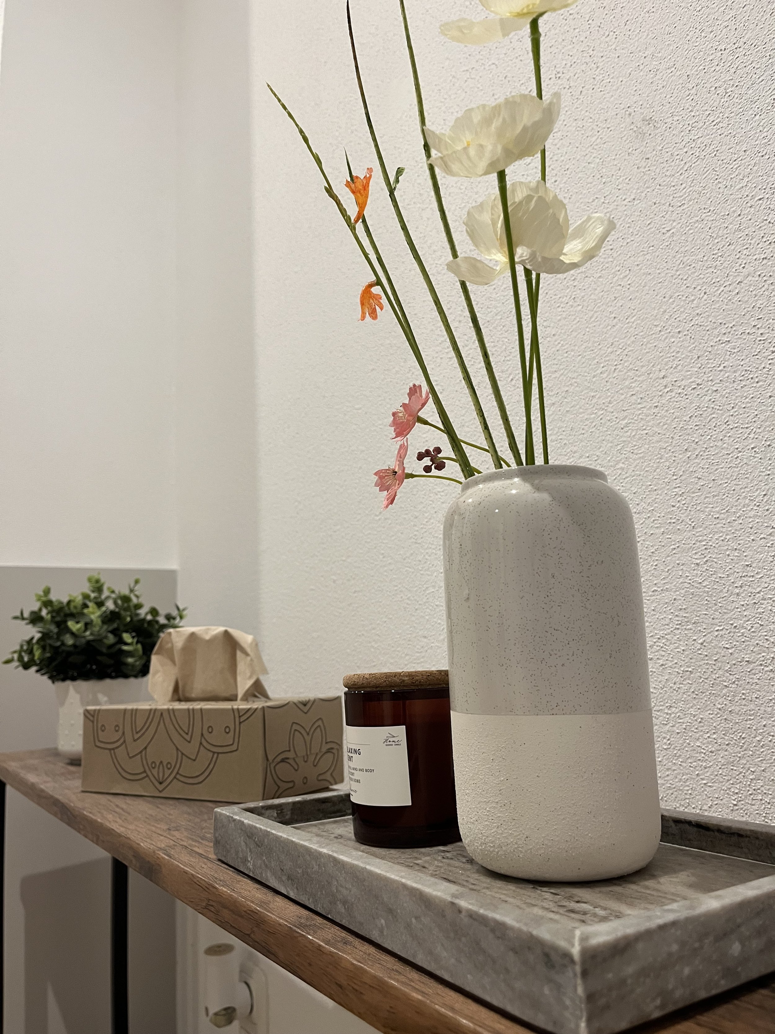 Vase mit weißen und gefärbten künstlichen Blumen auf einer Holz- und Steinterrasse, im Hintergrund eine kleine Topfpflanze und eine Geschenkbox.