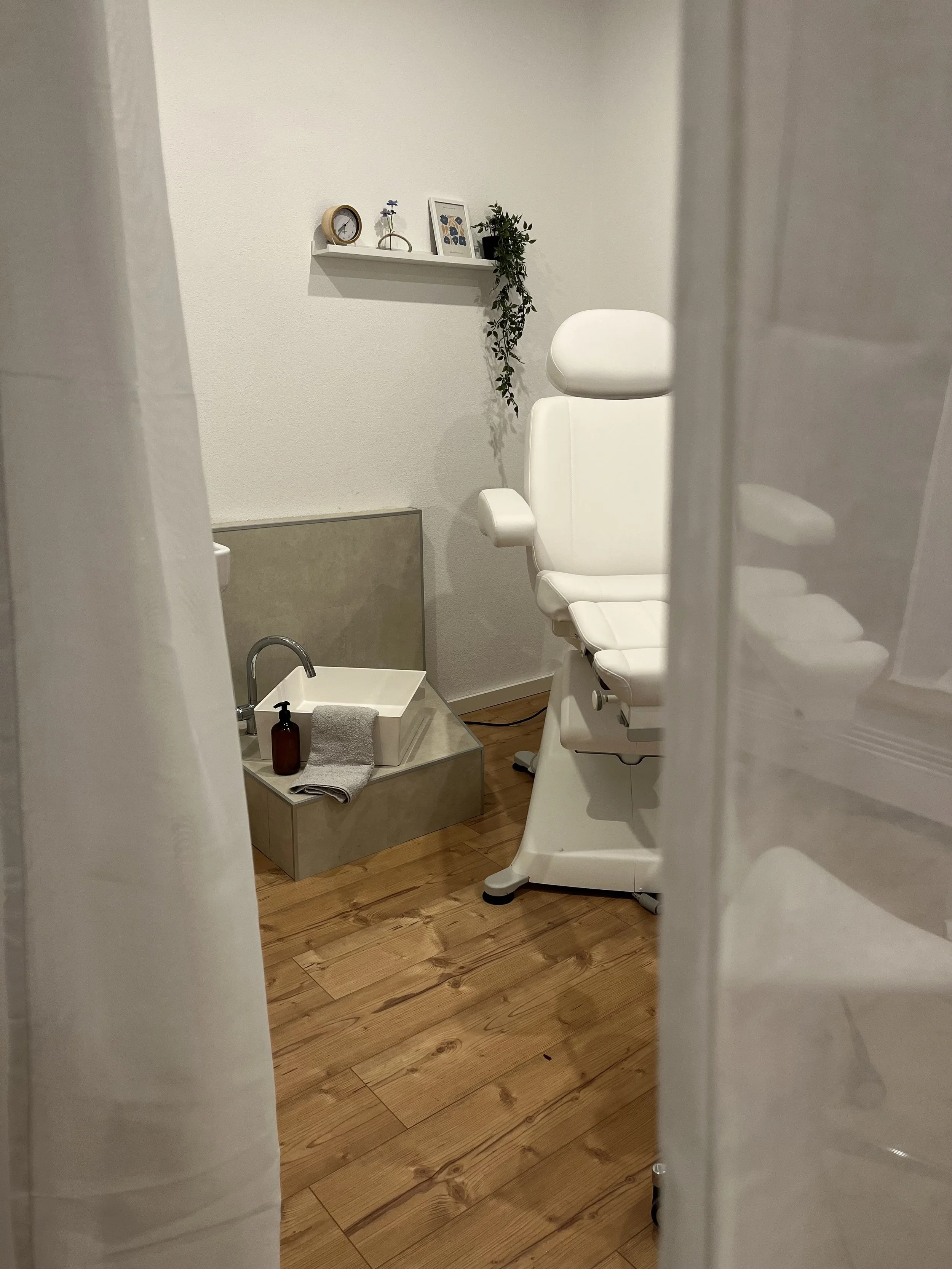 Ein weißer medizinischer Stuhl mit Kopfstütze in einem Behandlungsraum, daneben ein Fußbad mit Wasserhahn und Seife, an der Wand hängen eine kleine Uhr, Bilderrahmen und ein Pflanzenhänger, das Bild ist von einem Vorhang teilweise verdeckt.