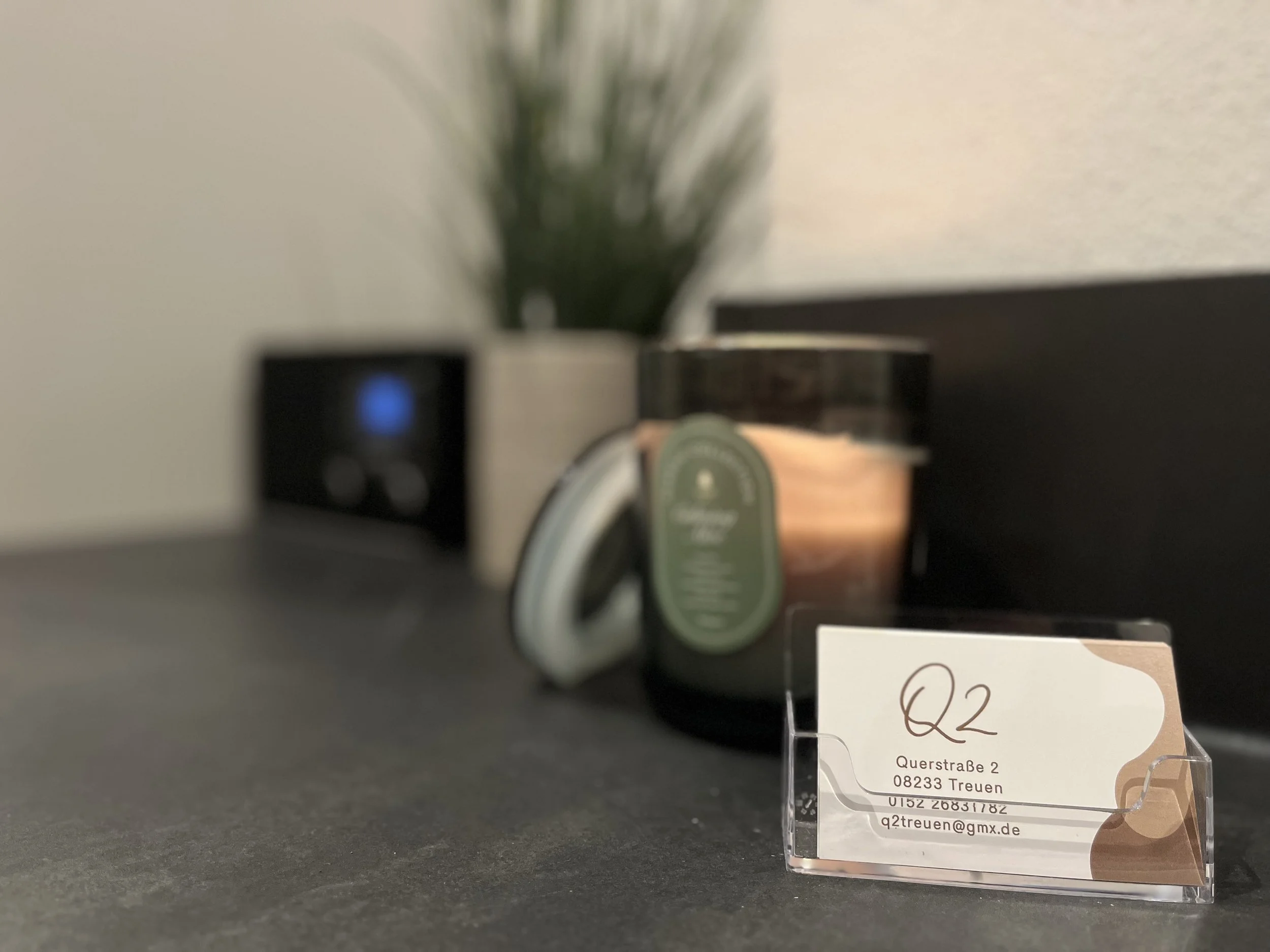 Geschäftskarte mit der Adresse Quersaße 2, 08233 Treuen, Deutschland, auf einem dunklen Tisch. Im Hintergrund sind ein Kerzenlicht, eine Pflanze in einem Topf, und ein elektronisches Gerät mit blauer Anzeige zu sehen.