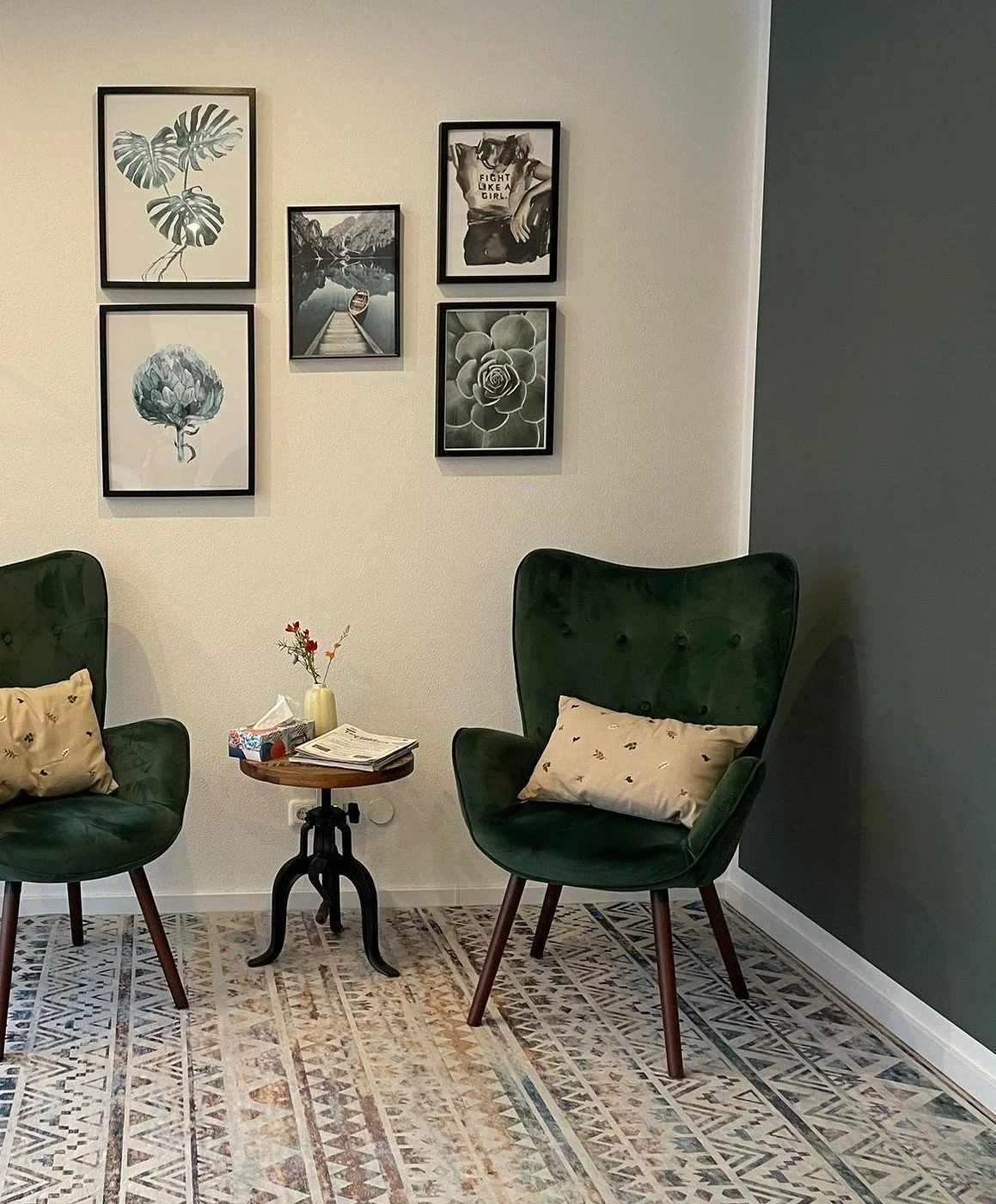 Zwei grüne Sessel mit Kissen, ein kleiner Beistelltisch mit eine Vase und Zeitschriften, Wanddekor mit sechs Rahmen, Teppichboden.