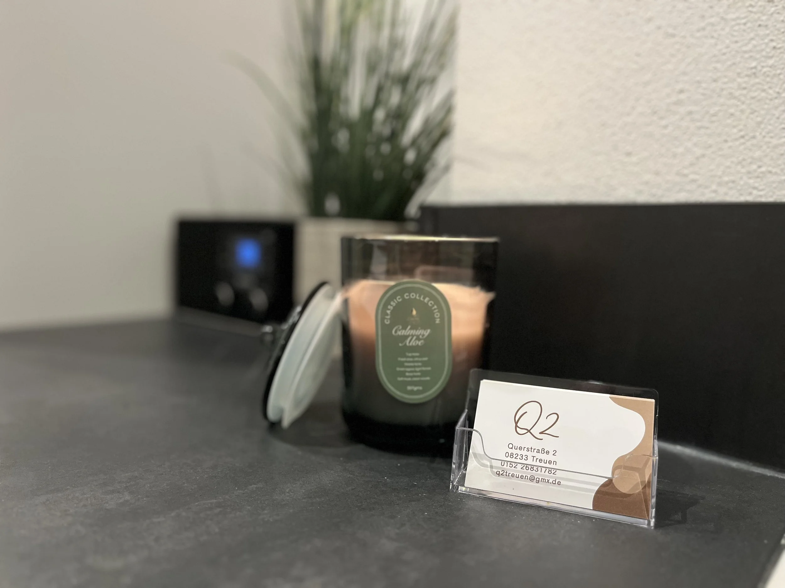 Ein schwarzer Kerzenleuchter mit einer Tasse Duftkerze mit grünem Etikett, ein Namensschild mit Adresse und E-Mail, im Hintergrund eine grüne Pflanze in einem weißen Topf auf einer schwarzen Oberfläche.