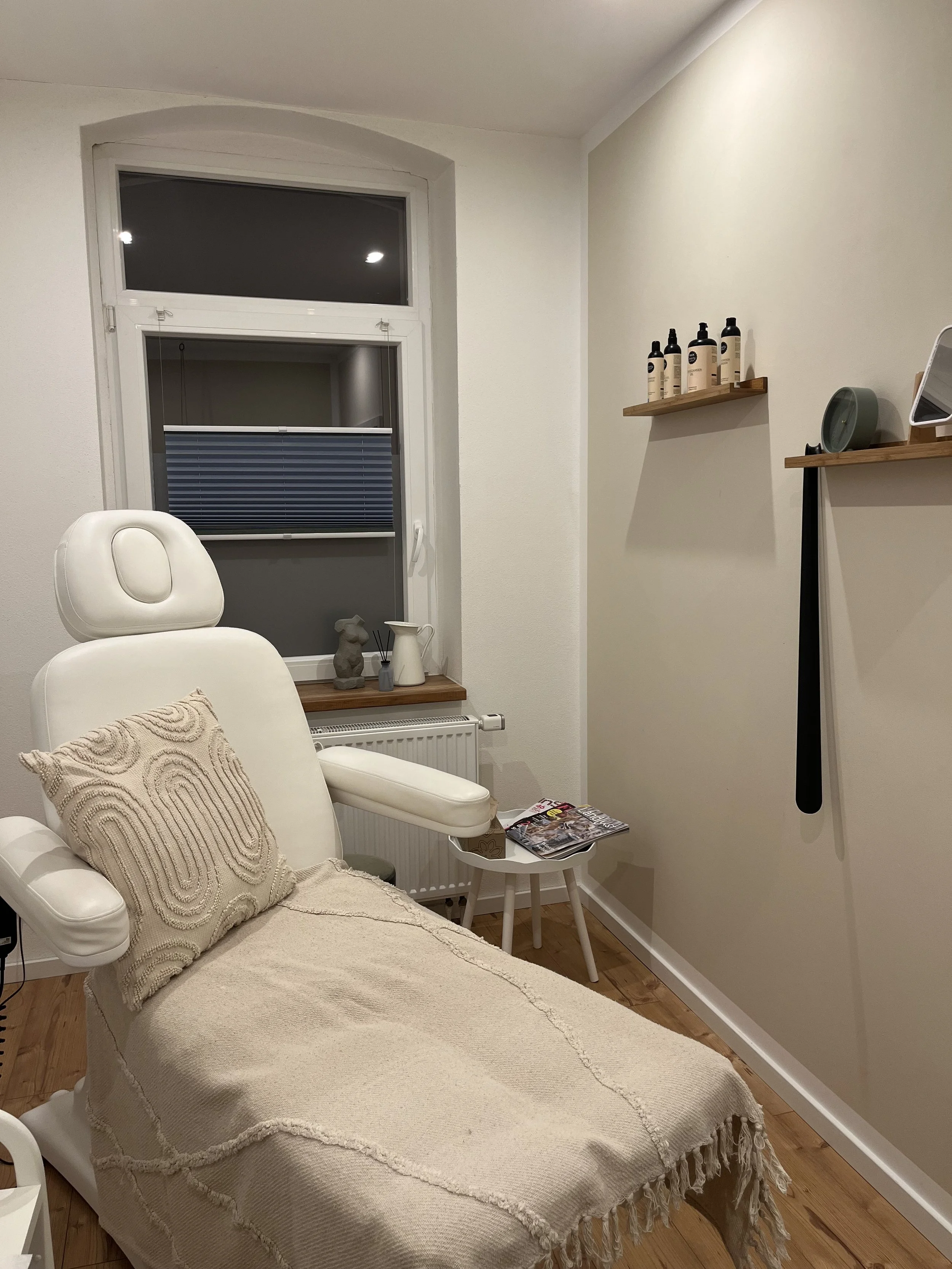 Ein Behandlungsraum mit einem weißen Behandlungstisch, einem Kissen, einem kleinen Beistelltisch mit Zeitschriften, einer Fensterbank mit Dekorationen, und Wandregalen mit Pflegeprodukten.