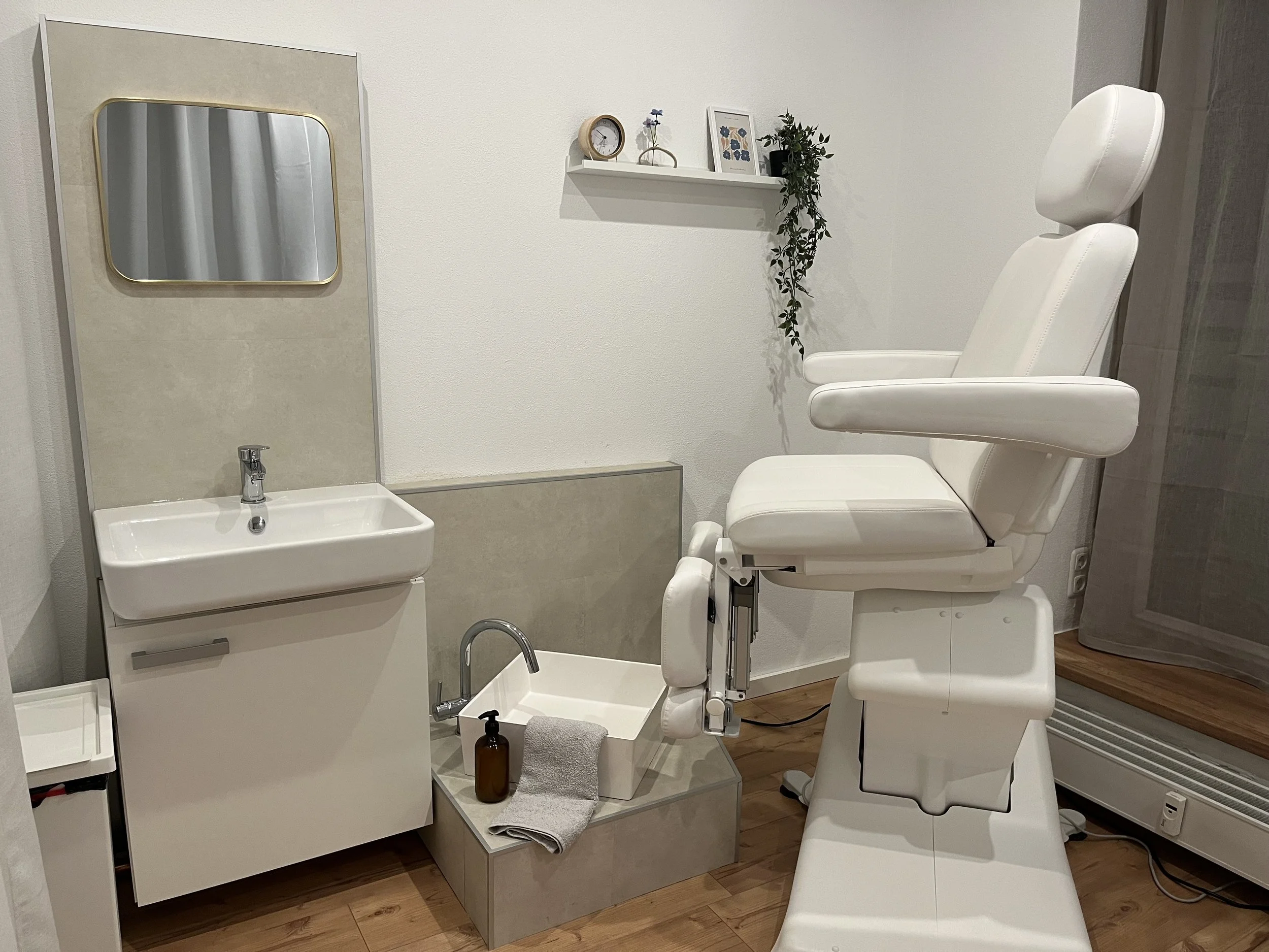 Ein Behandlungsraum in einer medizinischen oder therapeutischen Einrichtung mit einem weißen Stuhl, einer kleinen Waschgelegenheit, einem Spiegel und dekorativen Elementen auf einer Wandregal.