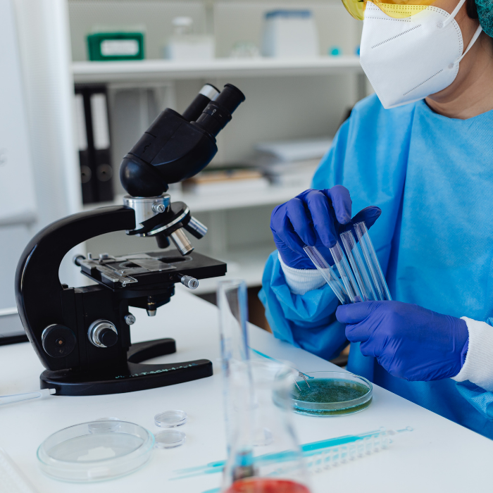A scientist in protective gear using test tubes in a laboratory with a microscope and lab equipment.