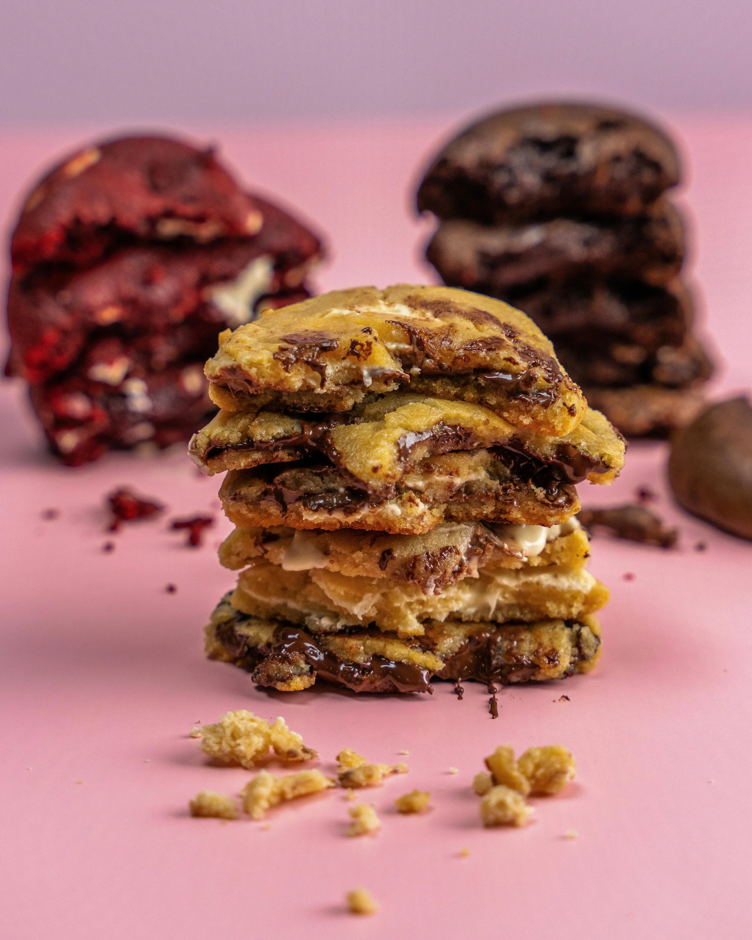 Stacked chocolate chip cookies with a pink background and scattered cookie crumbs, two stacks of cookies in the background.